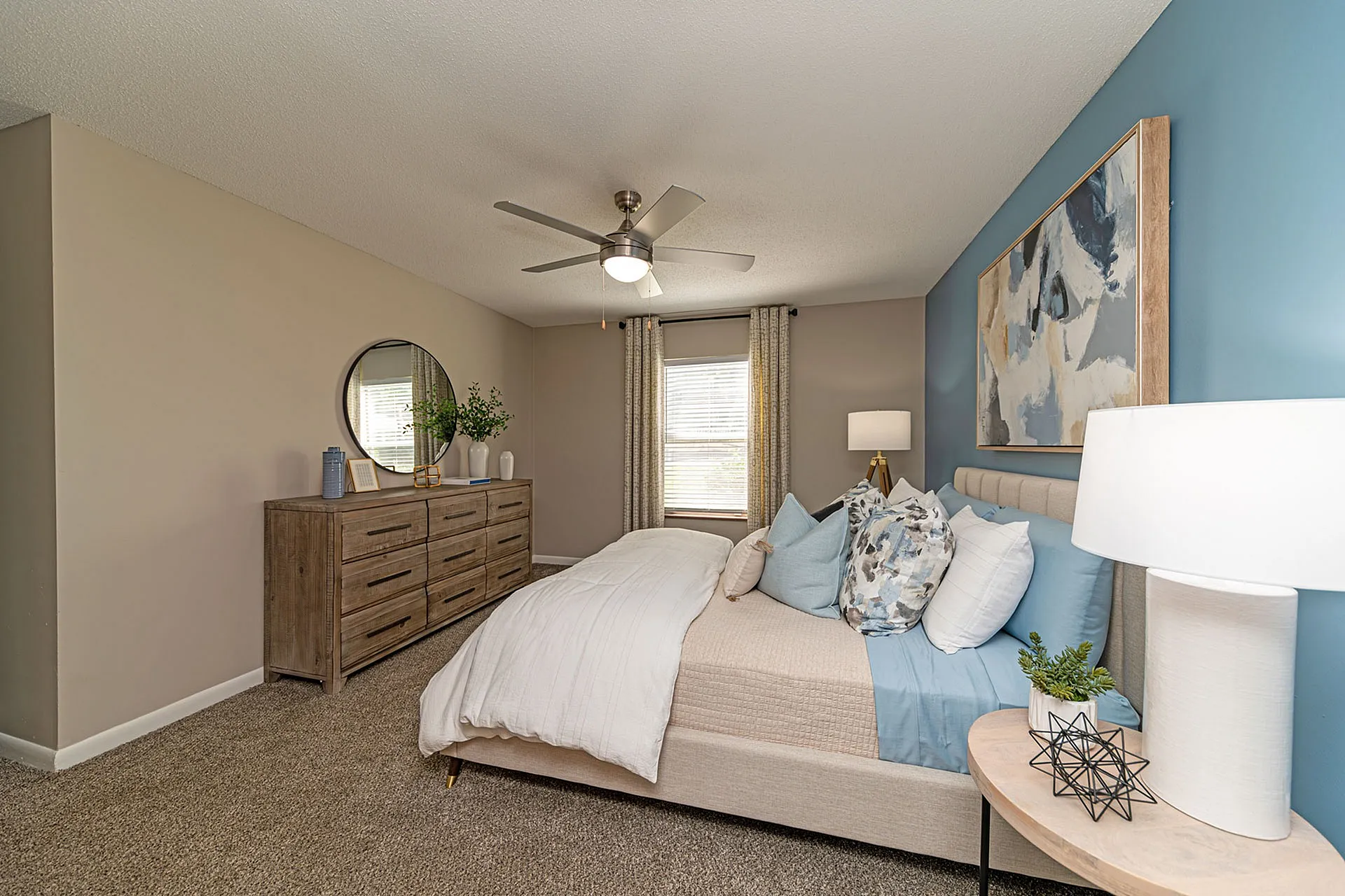 Elegant bedroom with modern decor, featuring a plush bed with stylish pillows, a wooden dresser with a round mirror, and calming blue accent wall.