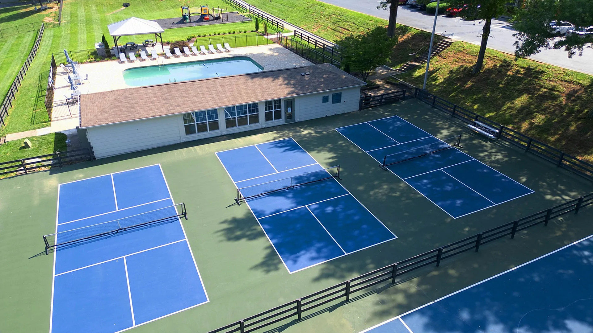 Vibrant aerial view of a community recreation area featuring multiple blue tennis courts and a sparkling swimming pool with lounge seating, surrounded by lush greenery.