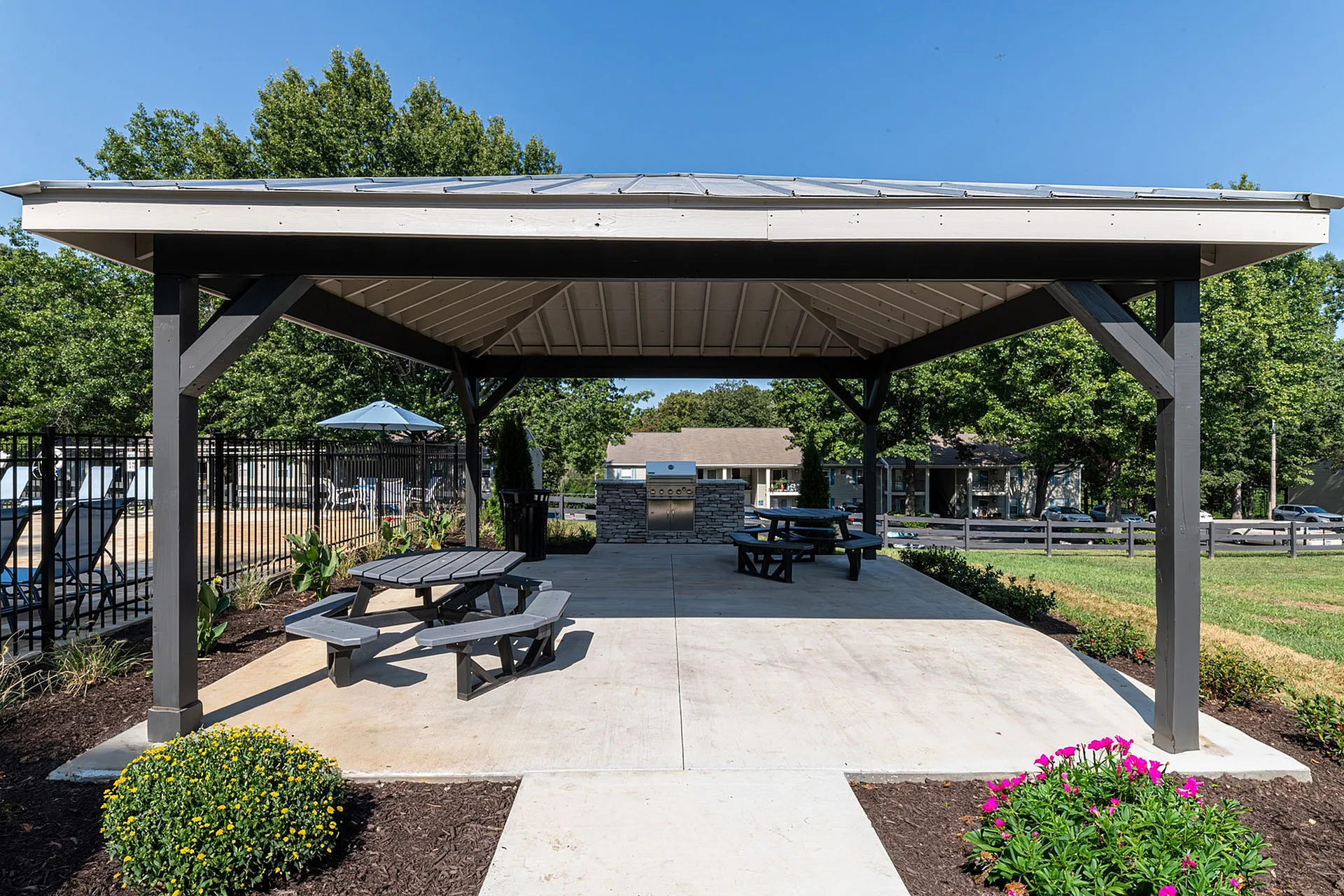 Charming outdoor pavilion with picnic tables and a stylish grill area, surrounded by lush greenery and vibrant flowers, perfect for community gatherings.
