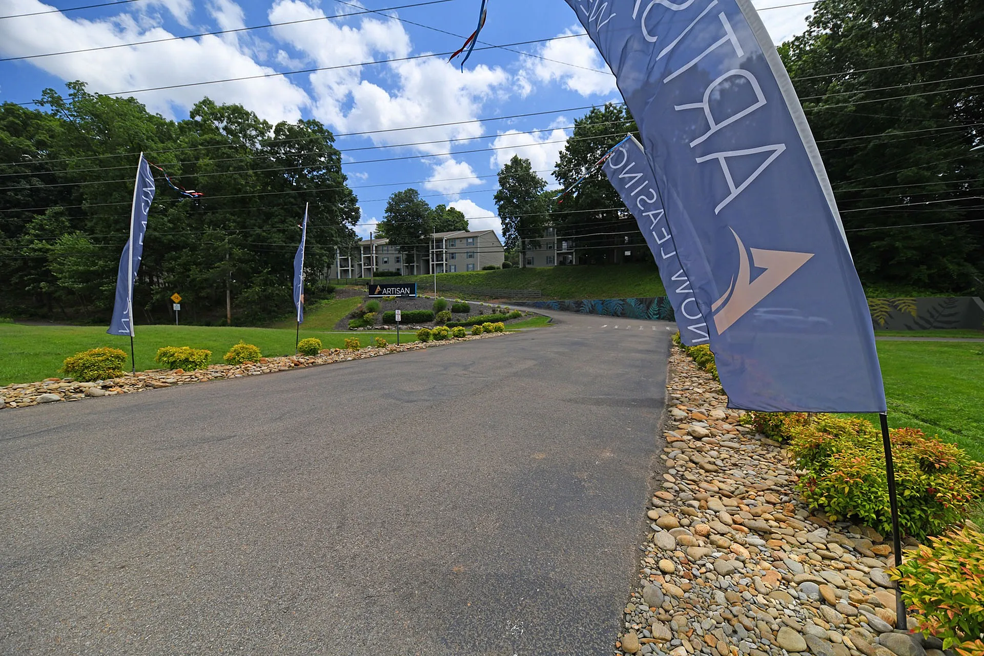 Inviting entrance to Artisan apartments, featuring lush greenery, vibrant flags, and a welcoming driveway on a sunny day.
