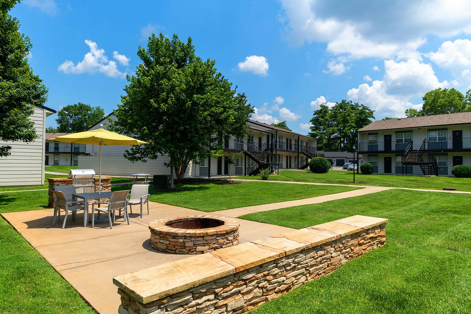 Charming apartment complex with inviting outdoor patio, lush green lawns, and a cozy fire pit under a sunny sky.