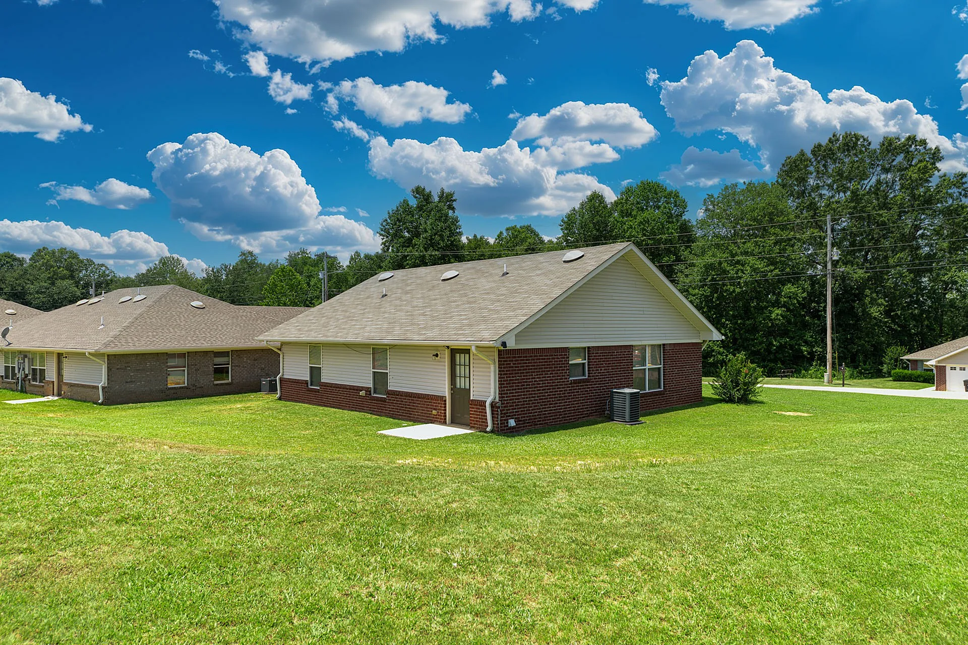 Charming single-story brick home with a spacious green lawn and picturesque countryside backdrop under a vibrant blue sky.