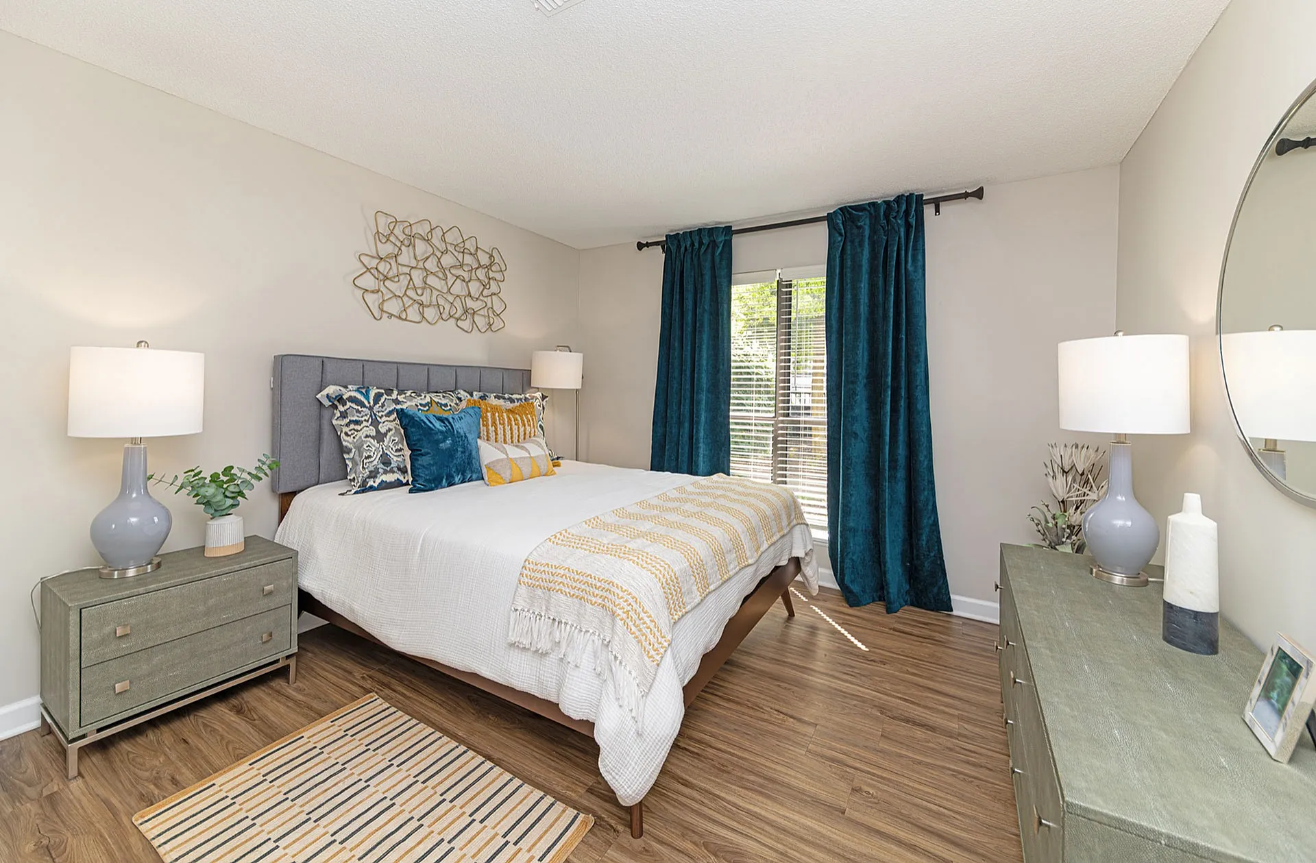 Inviting bedroom featuring plush decor with vibrant teal curtains, a luxurious bed set, and elegant lighting on wooden flooring.