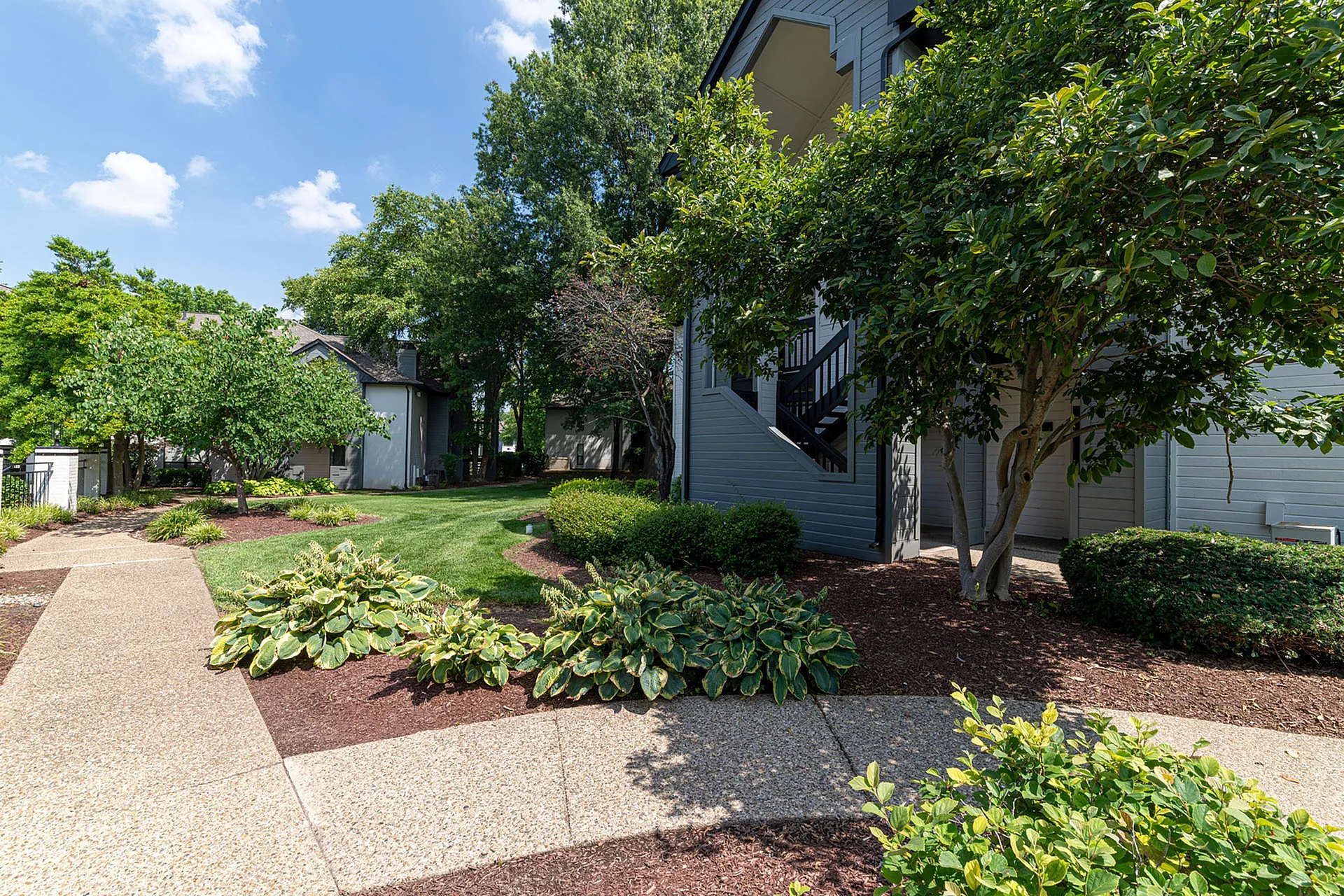 Charming apartment complex surrounded by lush greenery and well-manicured landscaping, featuring a welcoming pathway under a clear blue sky.