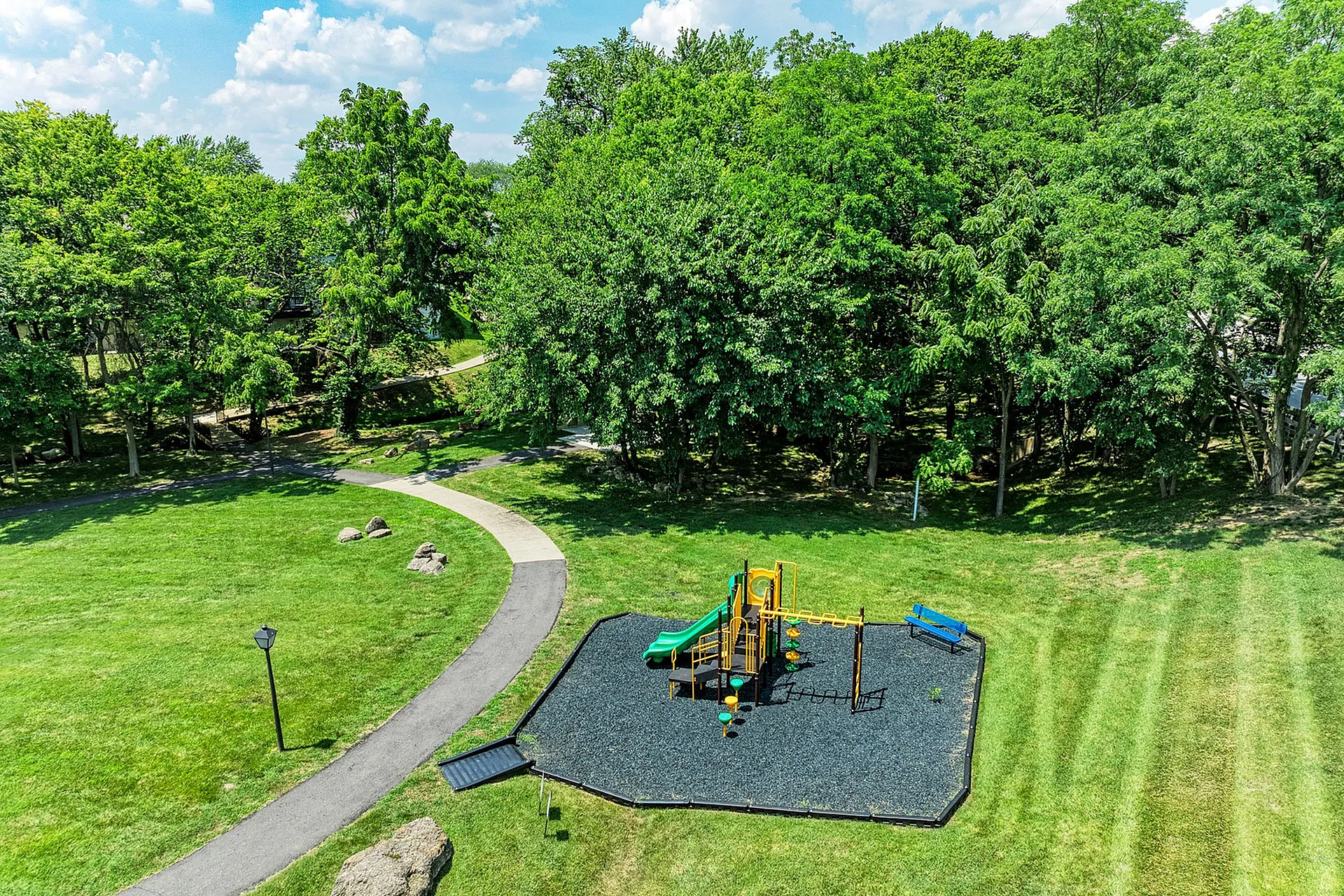 Inviting aerial view of a sunny park with lush greenery, featuring a vibrant playground surrounded by trees and open pathways.