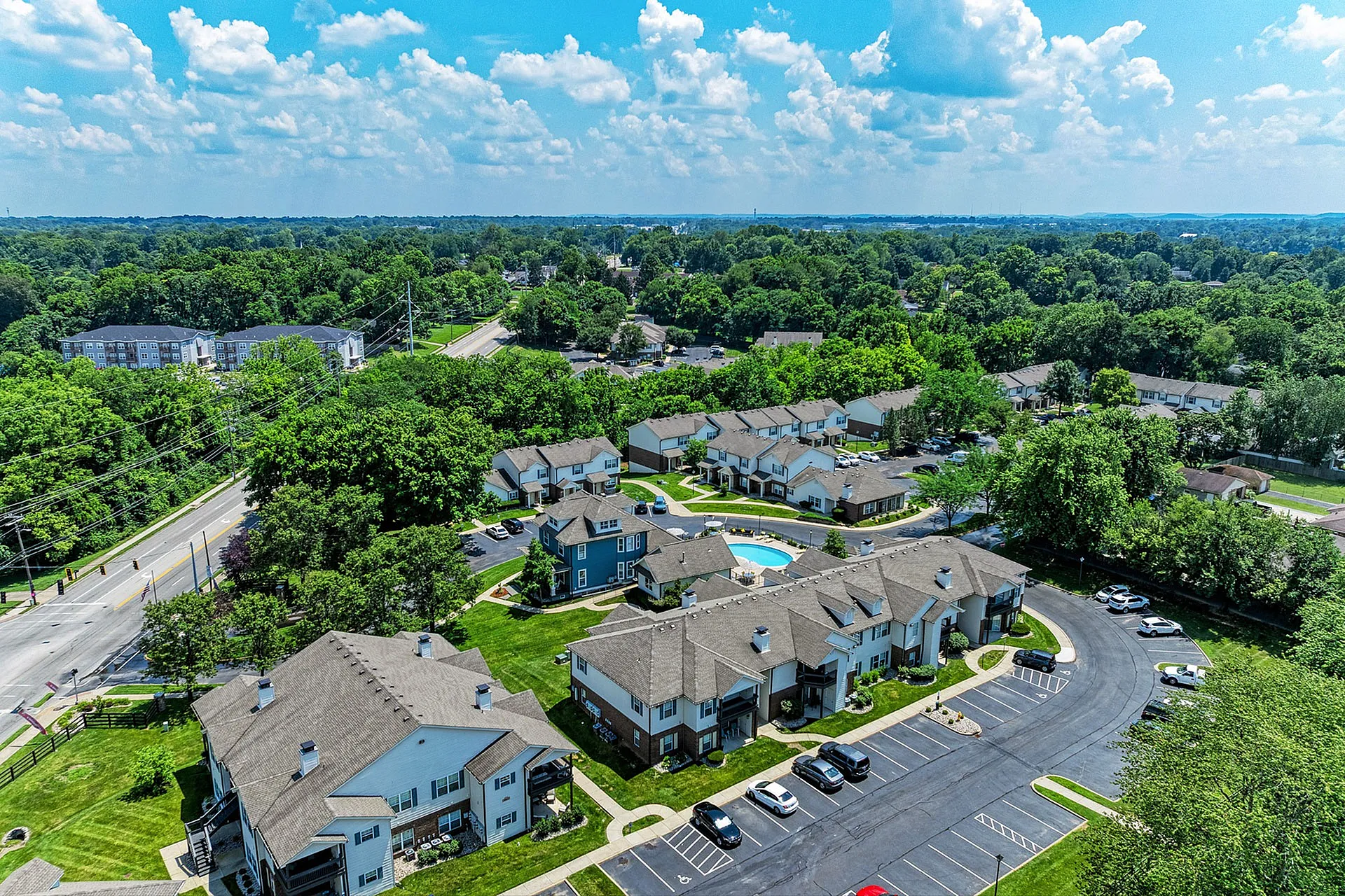Aerial view of a charming apartment complex surrounded by lush greenery, featuring well-maintained buildings, ample parking, and a central swimming pool under a bright blue sky.