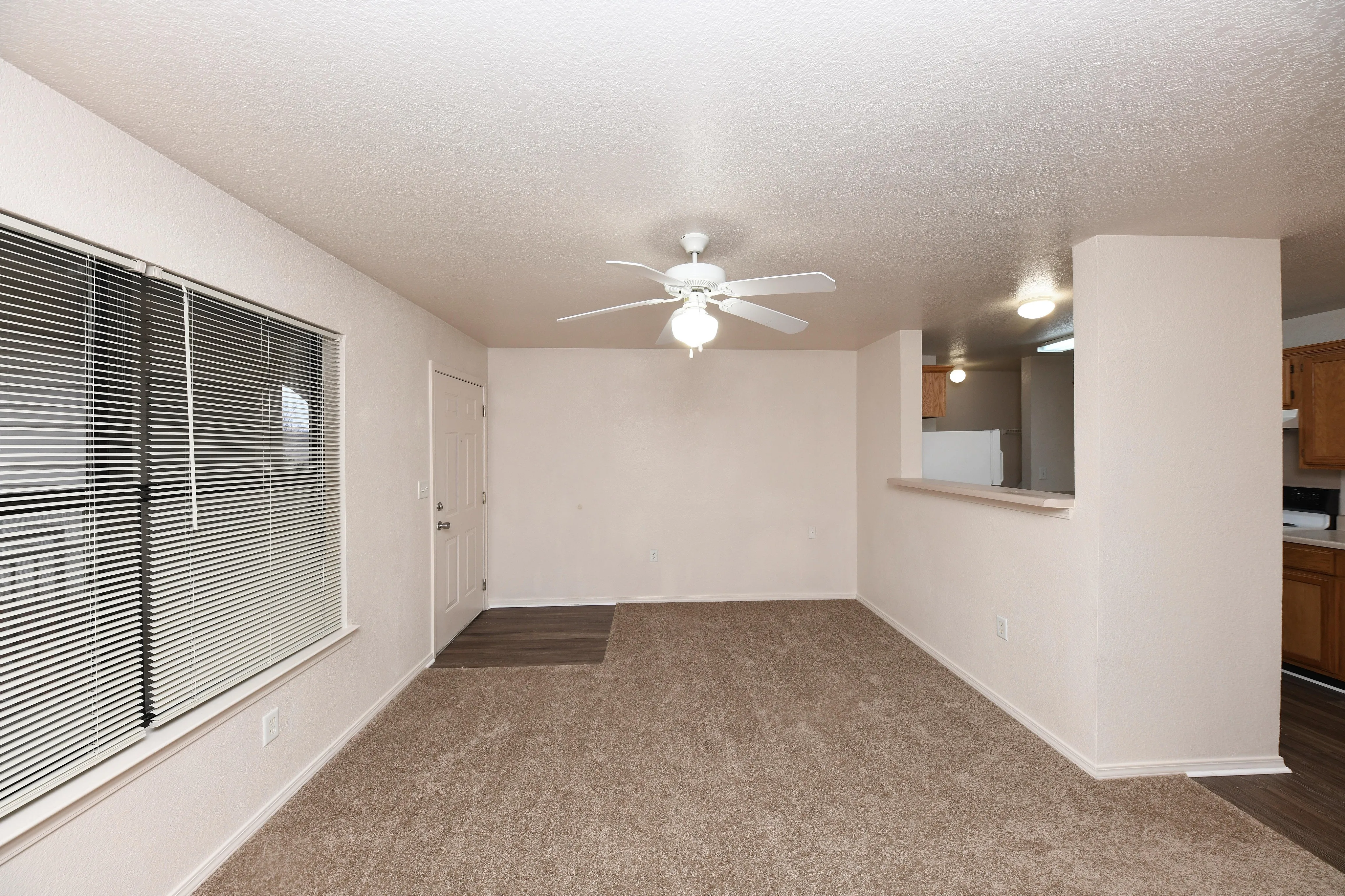 Bright and inviting living space featuring cozy carpet, ceiling fan, and ample natural light from large window blinds, adjacent to an open kitchen.