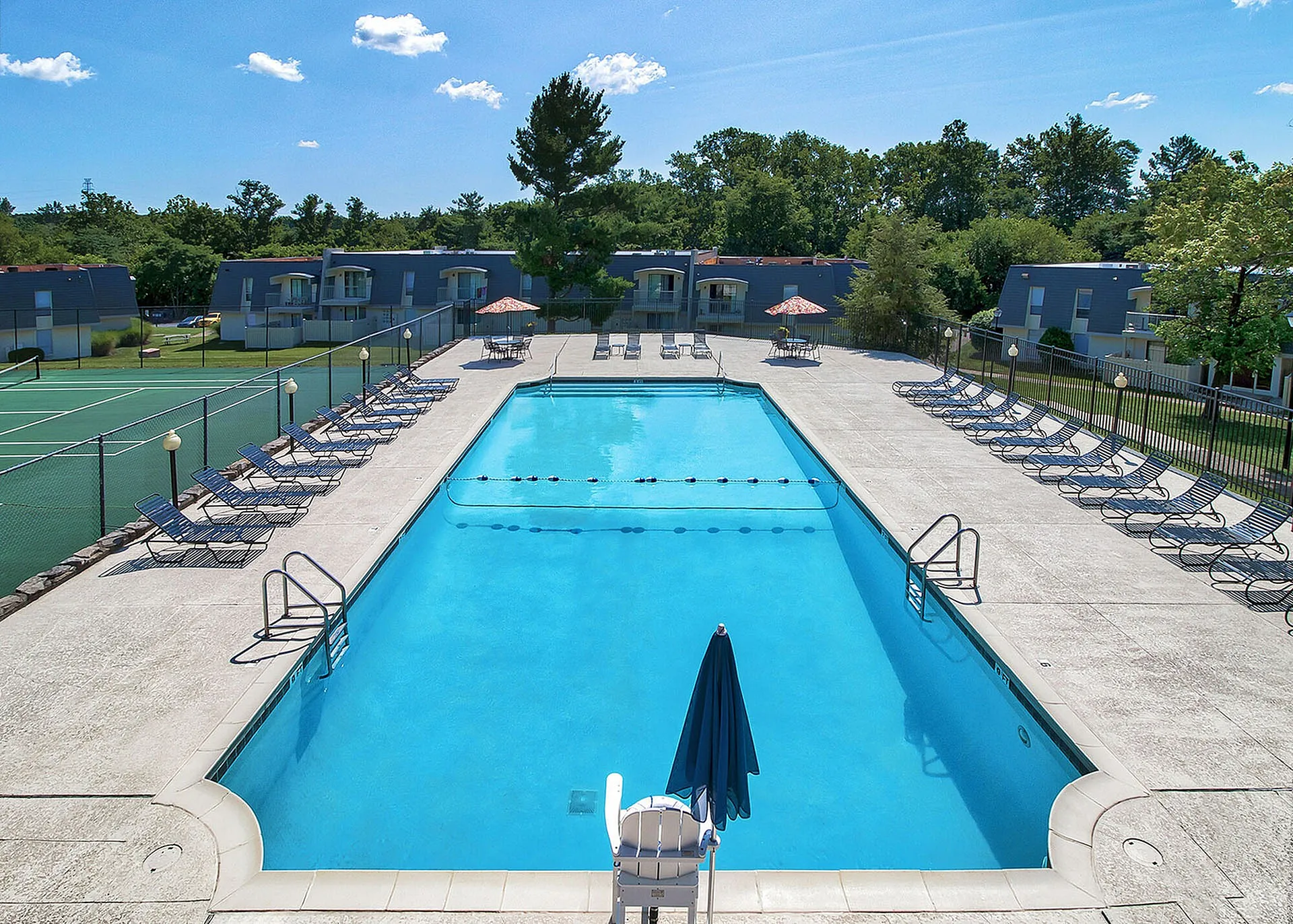 Luxurious apartment complex featuring a sparkling outdoor pool and tennis court, surrounded by lush greenery and modern architecture.
