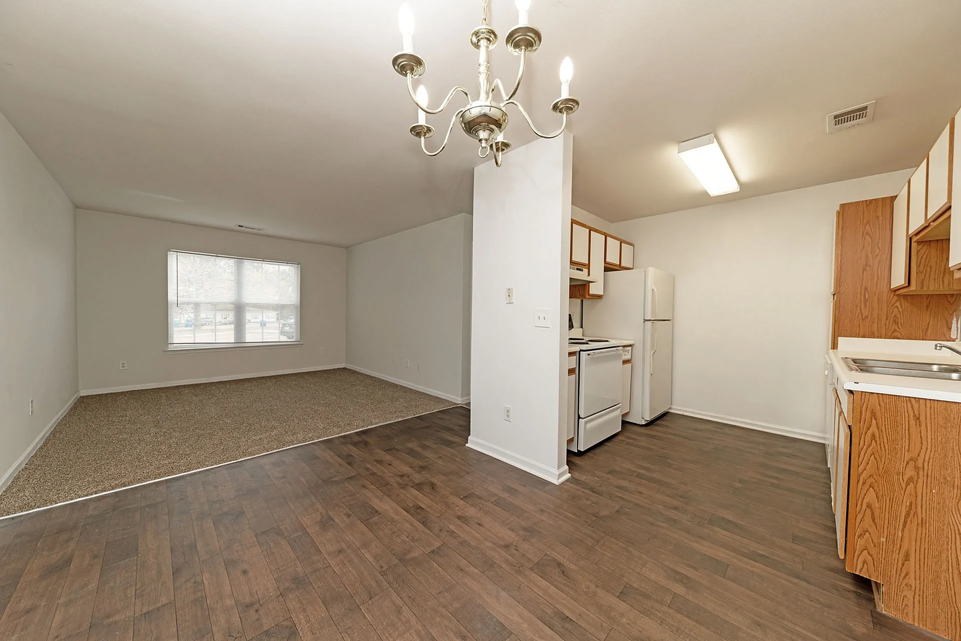 Inviting open plan apartment with elegant chandelier, cozy carpeted living space, and sleek hardwood floors leading to a modern kitchen.