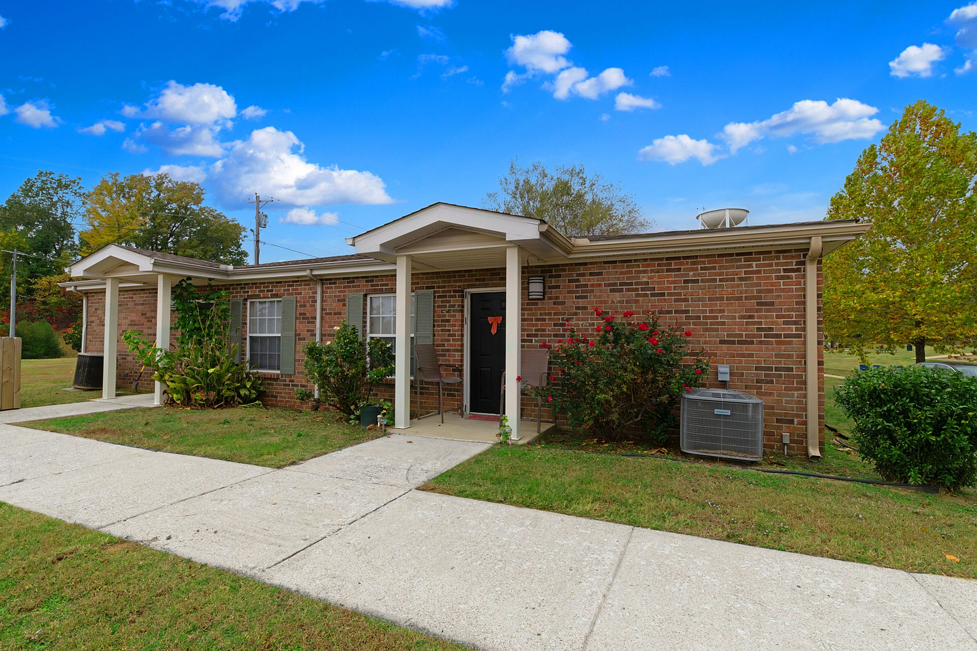Charming brick apartment exterior with lush landscaping and a welcoming front porch, set against a vibrant blue sky.