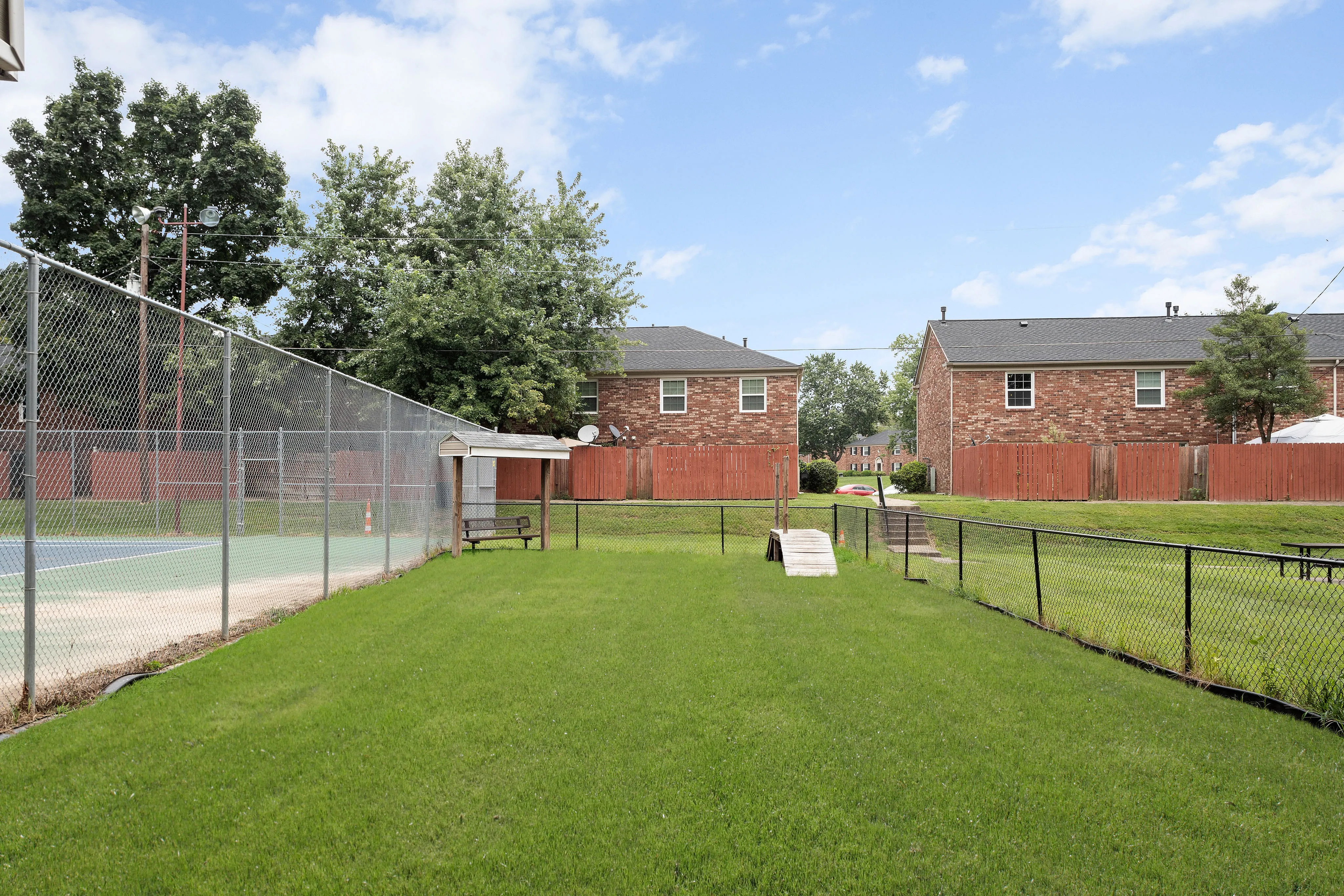 Inviting community area with lush green lawn and tennis court surrounded by charming brick houses under blue skies.