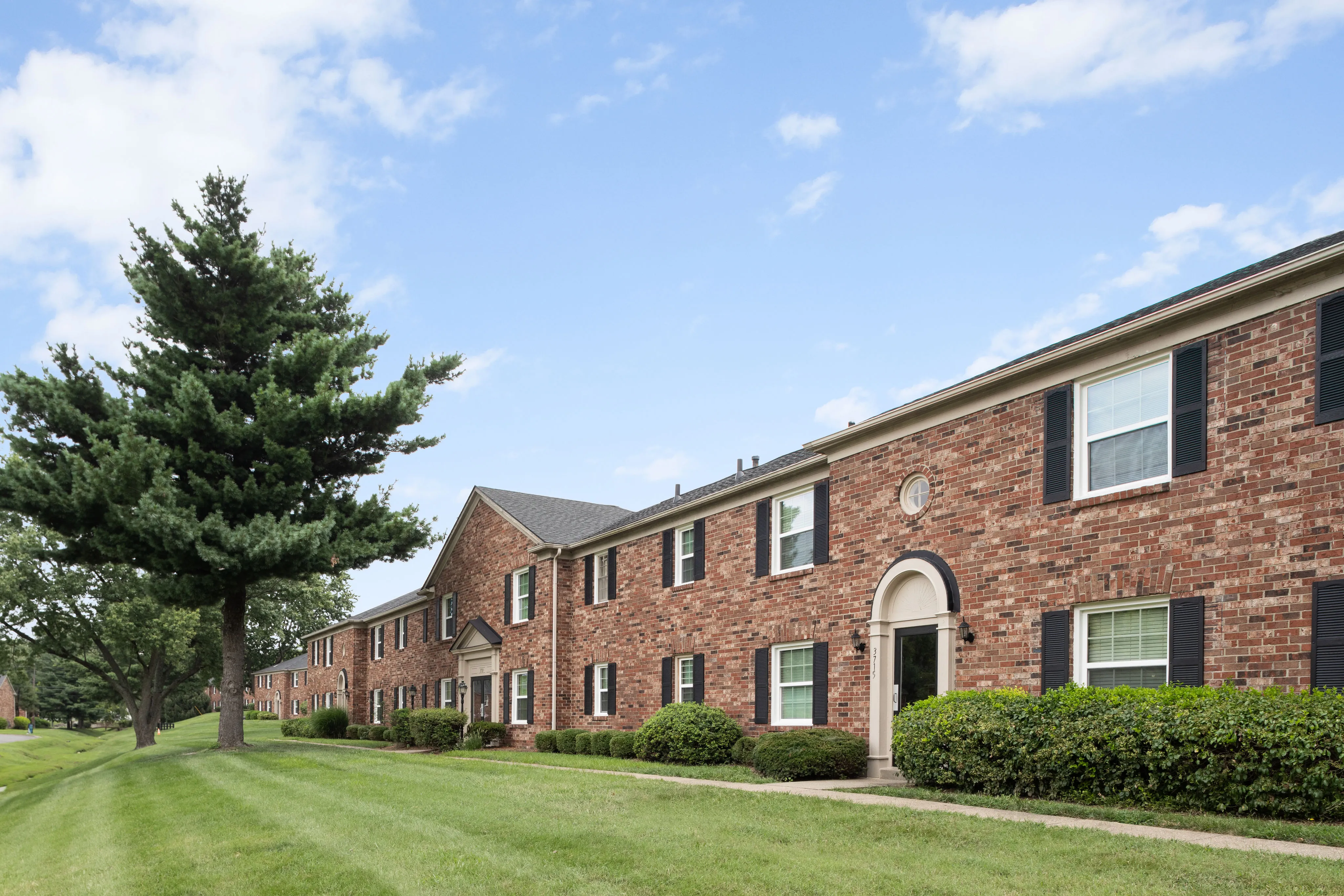 Charming brick apartment building with lush greenery and blue sky, offering a serene and inviting living environment.