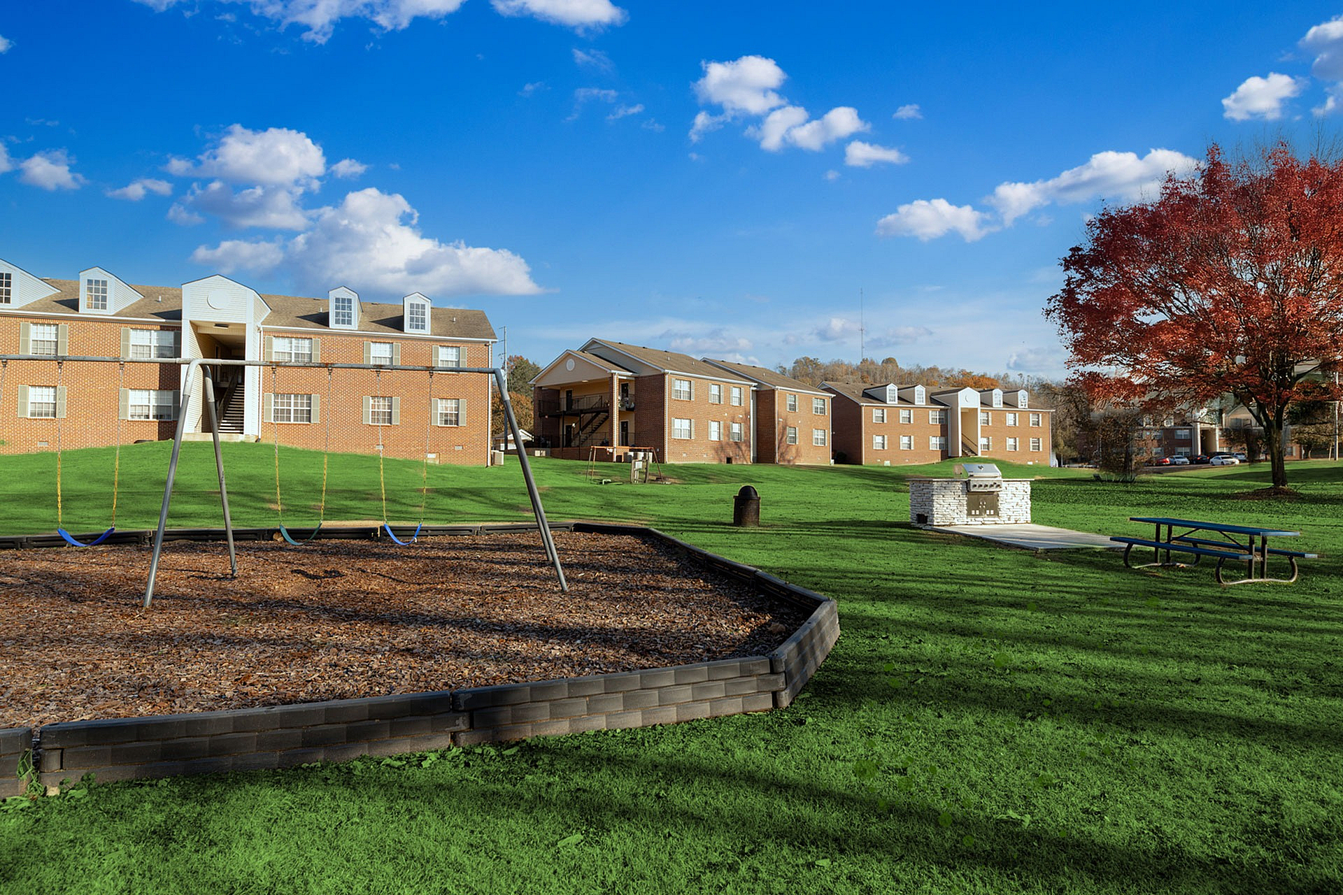 Charming residential complex with brick buildings, lush green lawns, playground, picnic tables, and serene blue sky.
