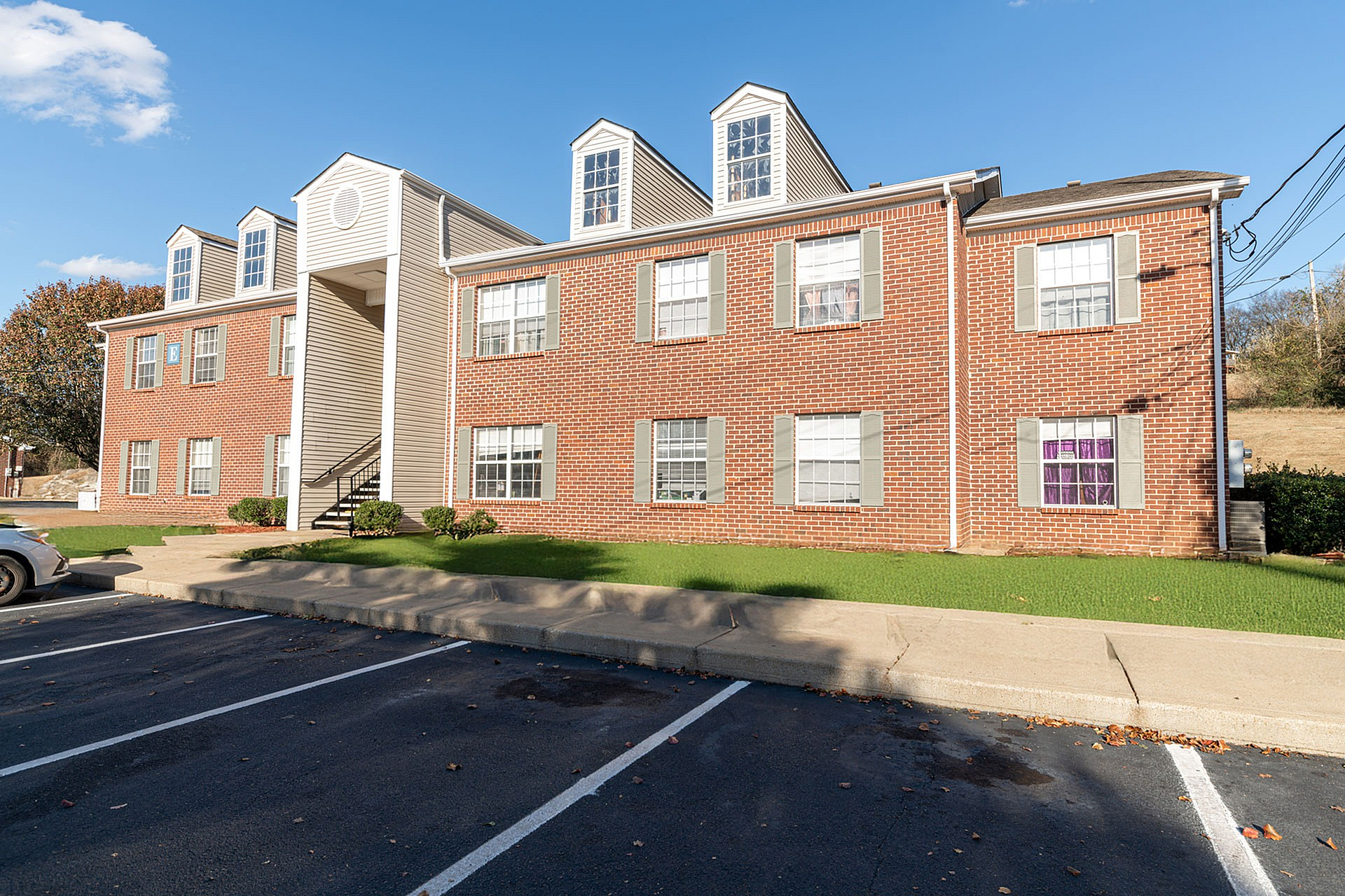 Charming brick apartment building with classic architecture, featuring manicured lawns and ample parking under a bright blue sky.