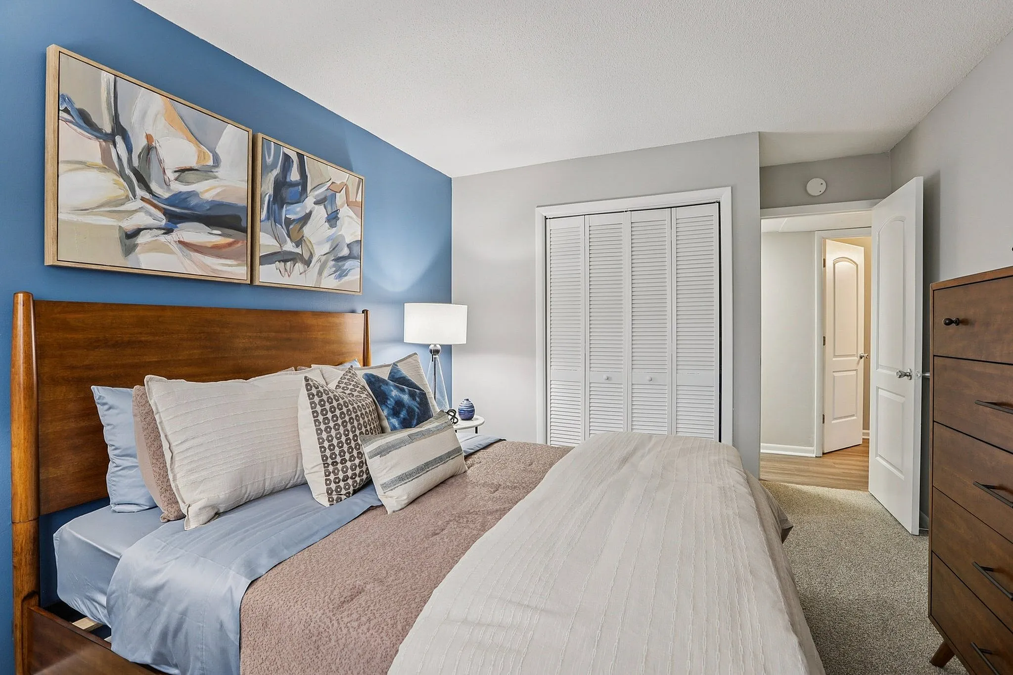 Inviting bedroom featuring a cozy wooden bed, bright accent pillows, modern artwork, and a calming blue accent wall.