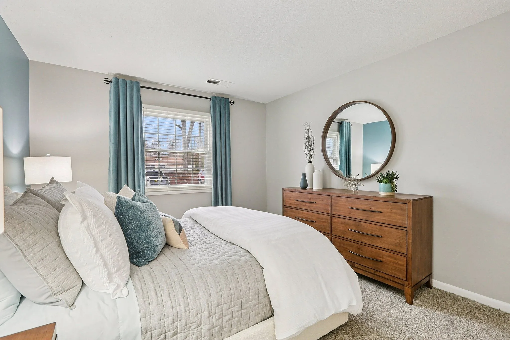 Inviting bedroom with plush bedding, elegant wooden dresser, and a large circular mirror, bathed in natural light from a spacious window with teal curtains.