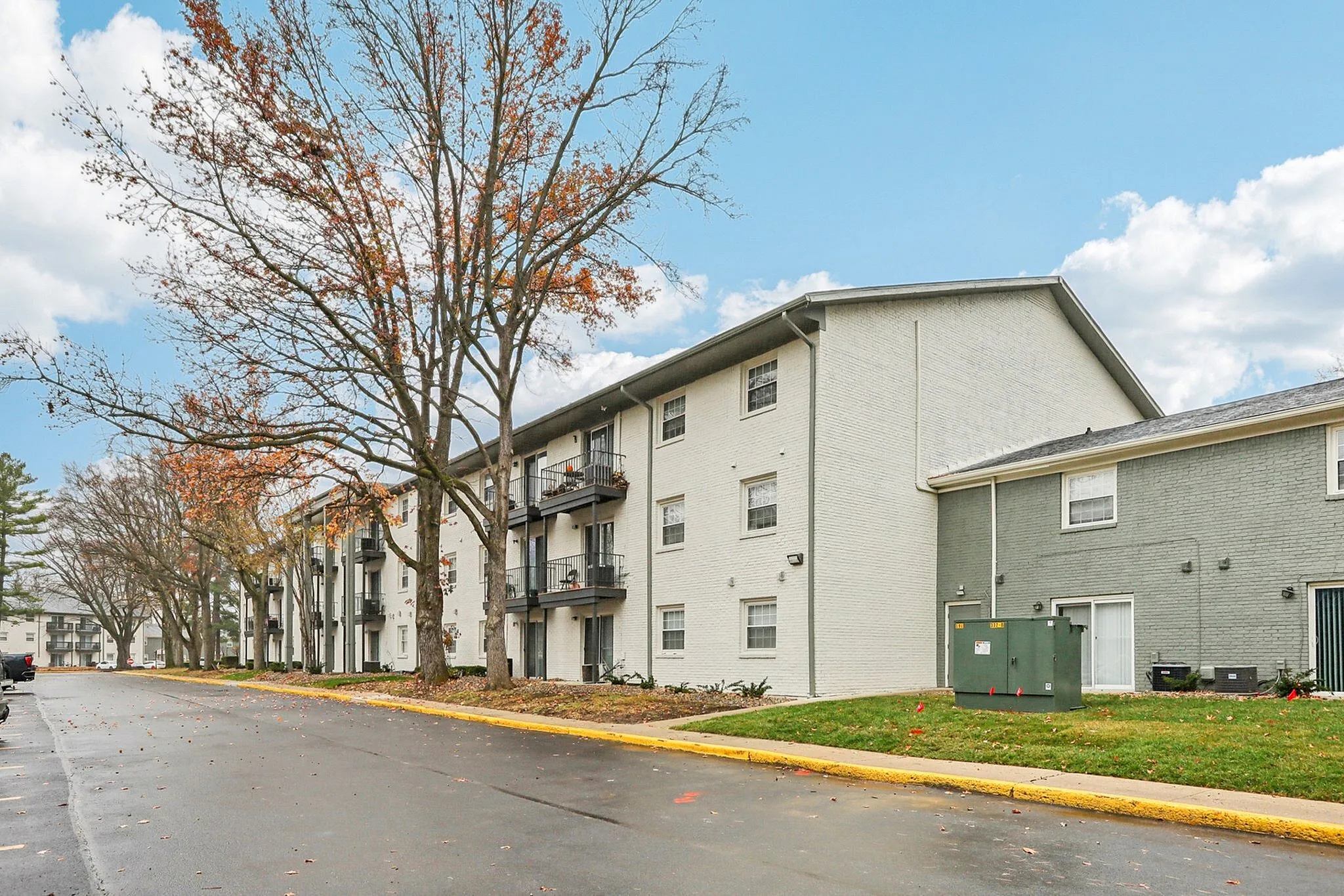 Charming apartment complex exterior featuring modern balconies, surrounded by lush trees and wide pathways, under a bright blue sky.