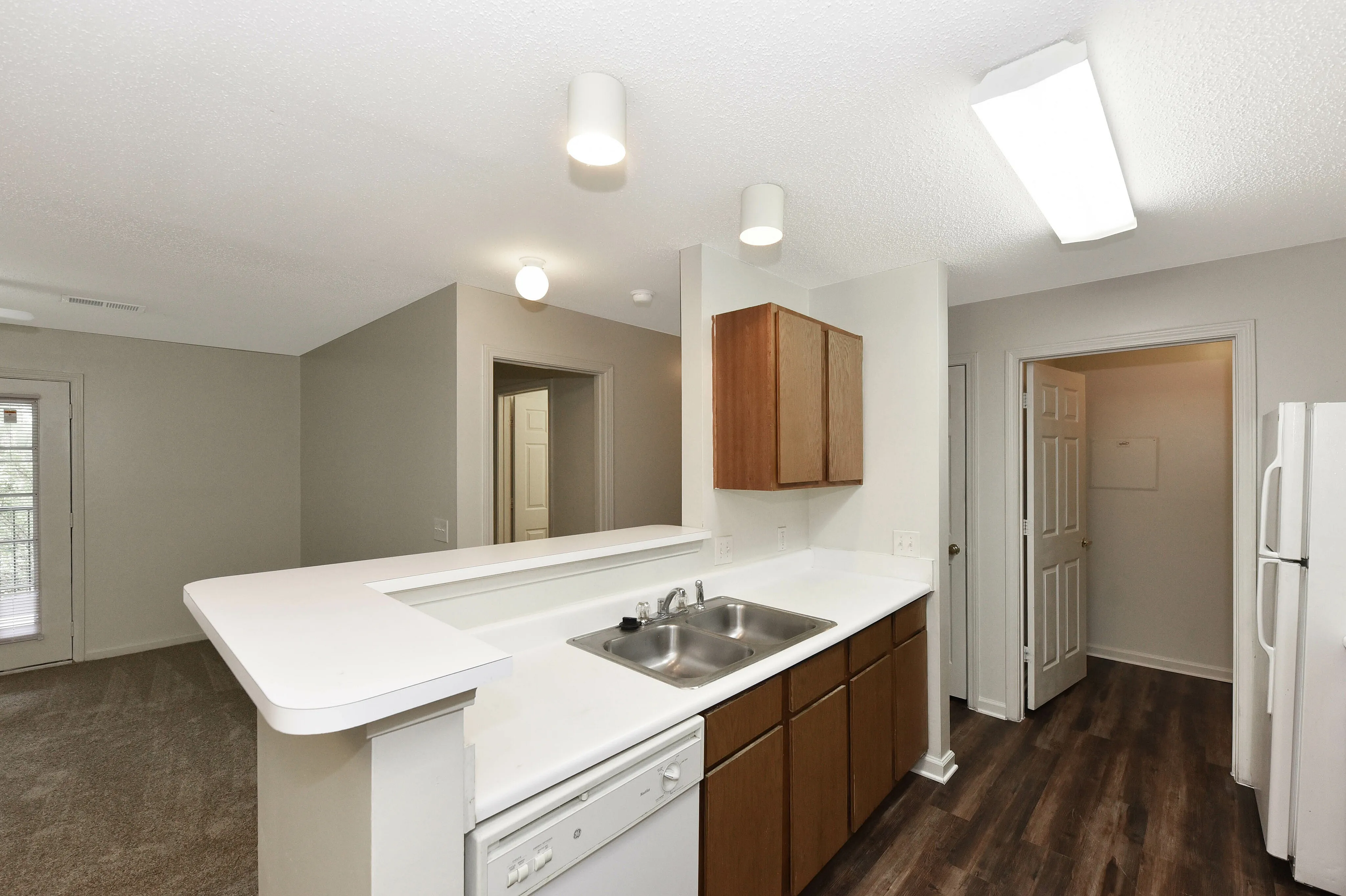 Modern kitchen with sleek countertops, stainless steel sink, and stylish cabinetry, open to a cozy living area with carpet flooring and natural light.
