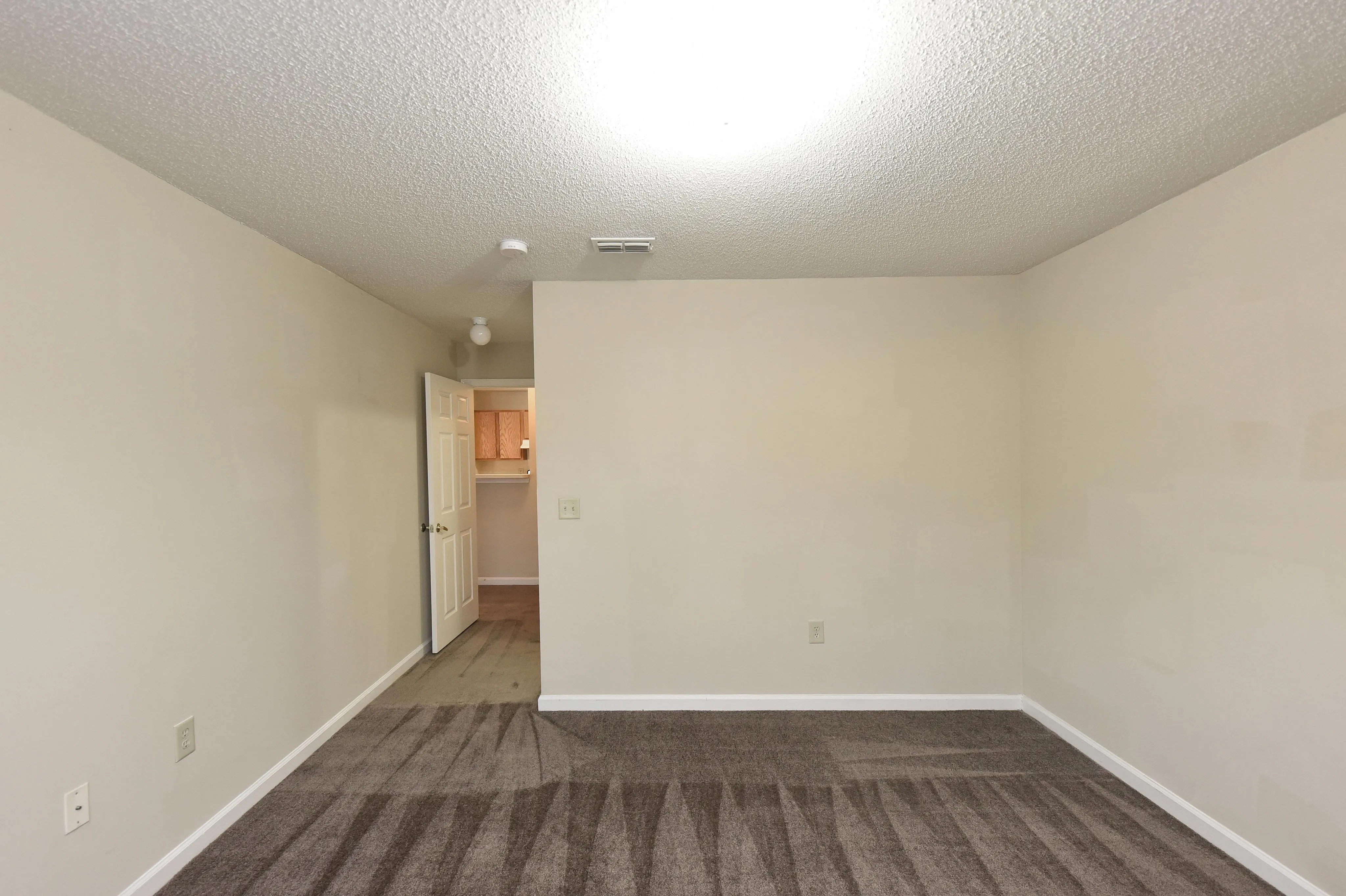 Inviting empty room with plush carpet and neutral walls, leading to a cozy hallway with a glimpse of the kitchen area.