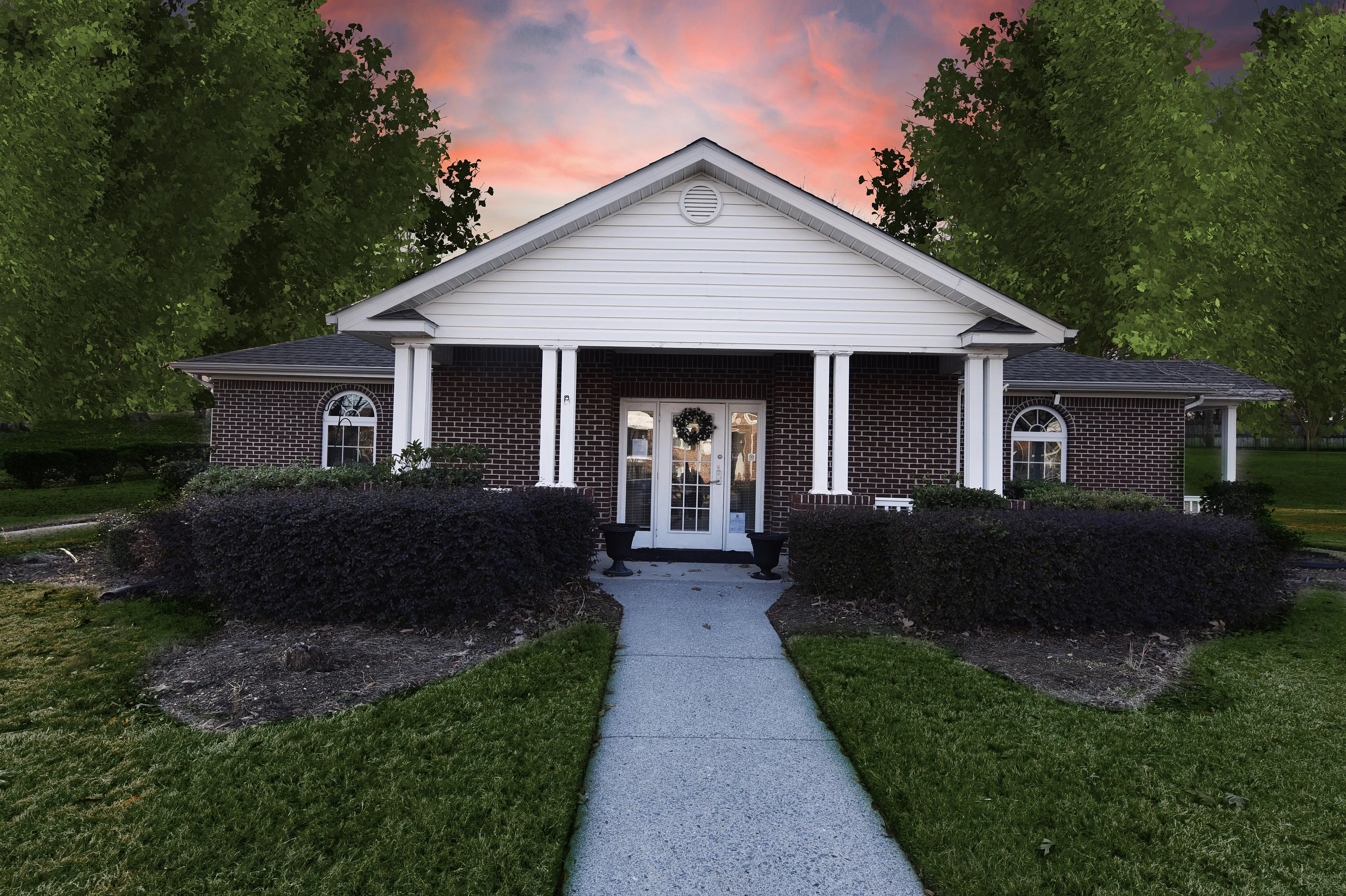 sCharming single-story brick home with classic columns, surrounded by lush greenery and a vibrant sunset backdrop.