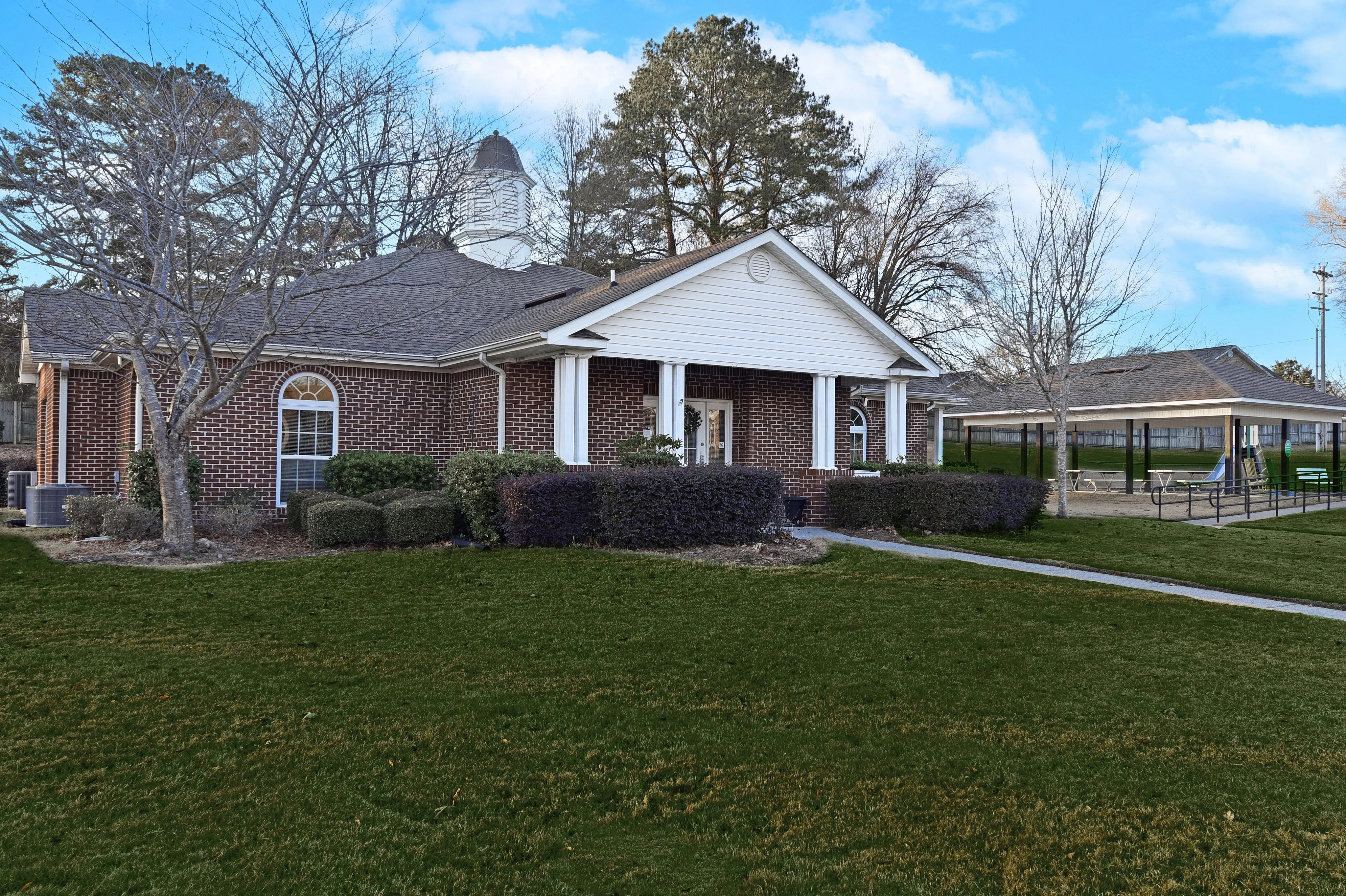 Charming brick building with a classic white portico, surrounded by lush greenery under a bright blue sky.