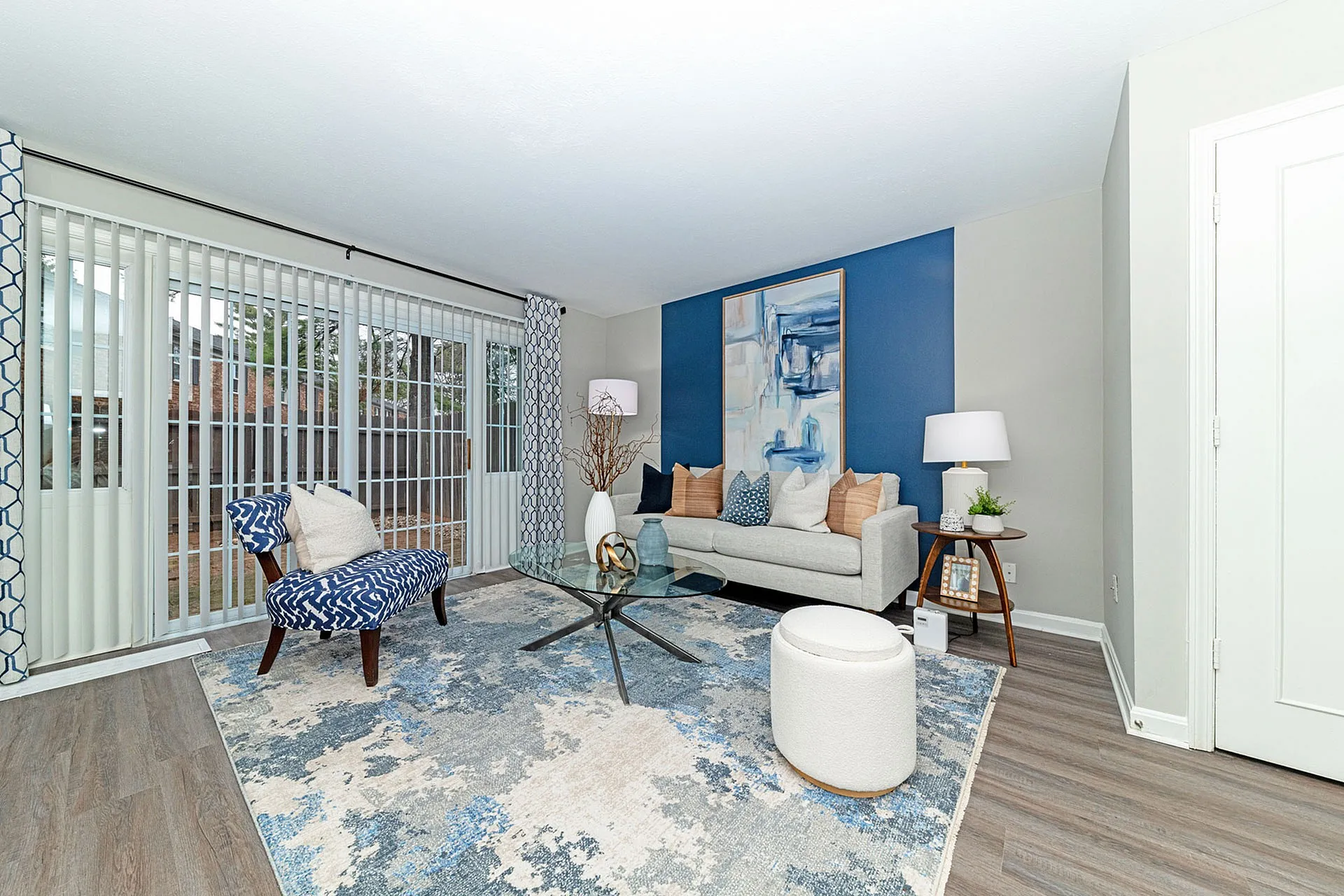 Bright and modern living room featuring large windows with vertical blinds, elegant decor, and a cozy seating area on a stylish patterned rug.