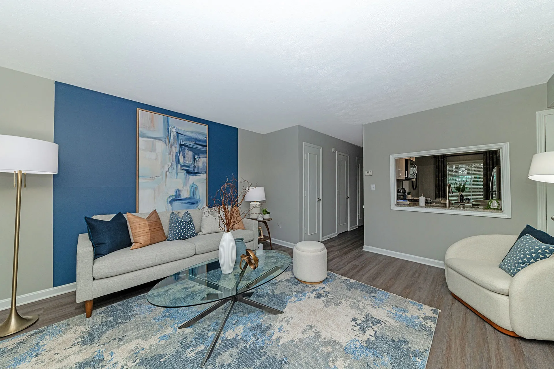 Chic living room with modern decor featuring a sleek glass coffee table, cozy sofa with stylish cushions, vibrant blue accent wall, and a window view into the kitchen.