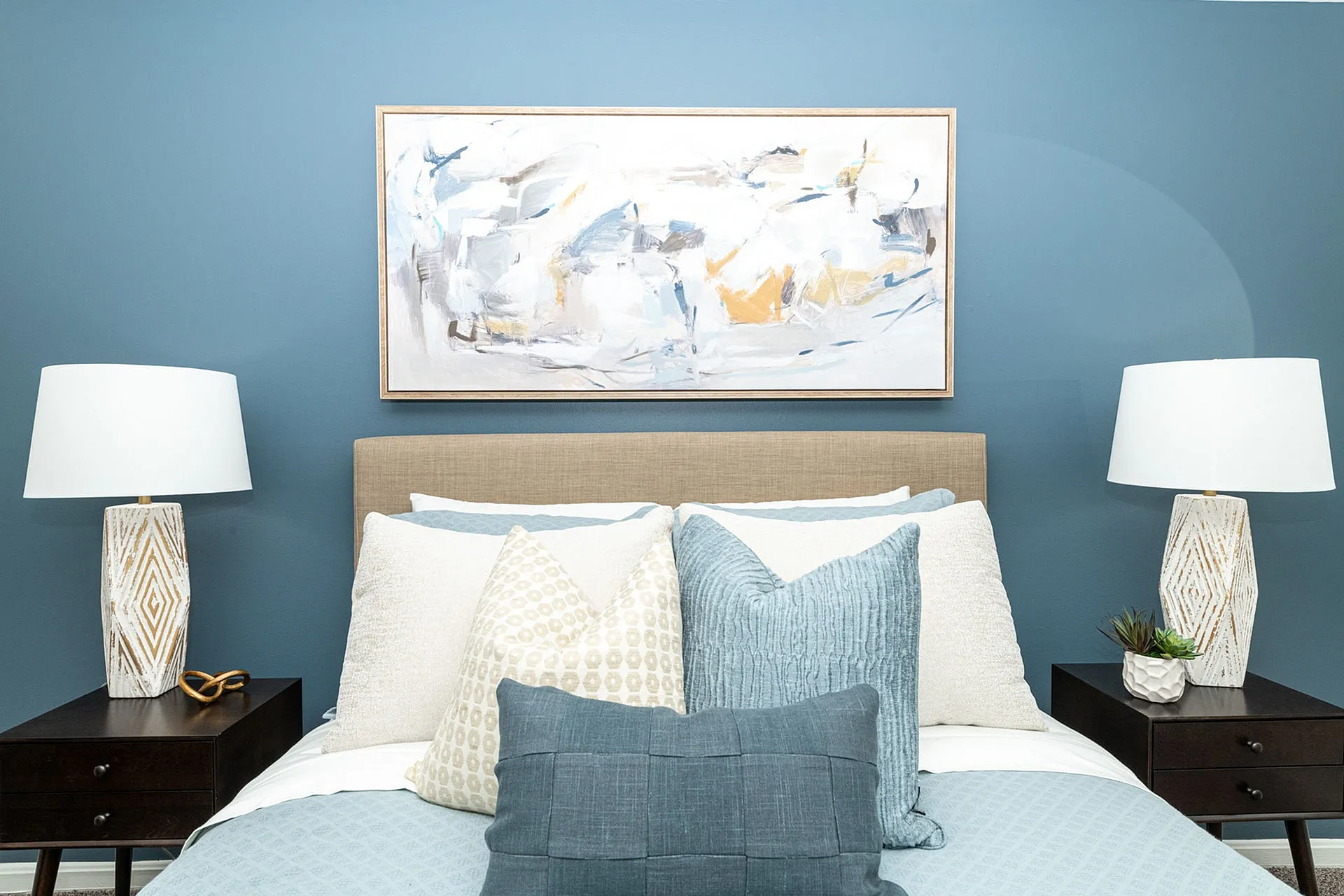 Elegant bedroom with modern decor featuring a plush bed, stylish abstract artwork, and chic bedside lamps against a serene blue wall.