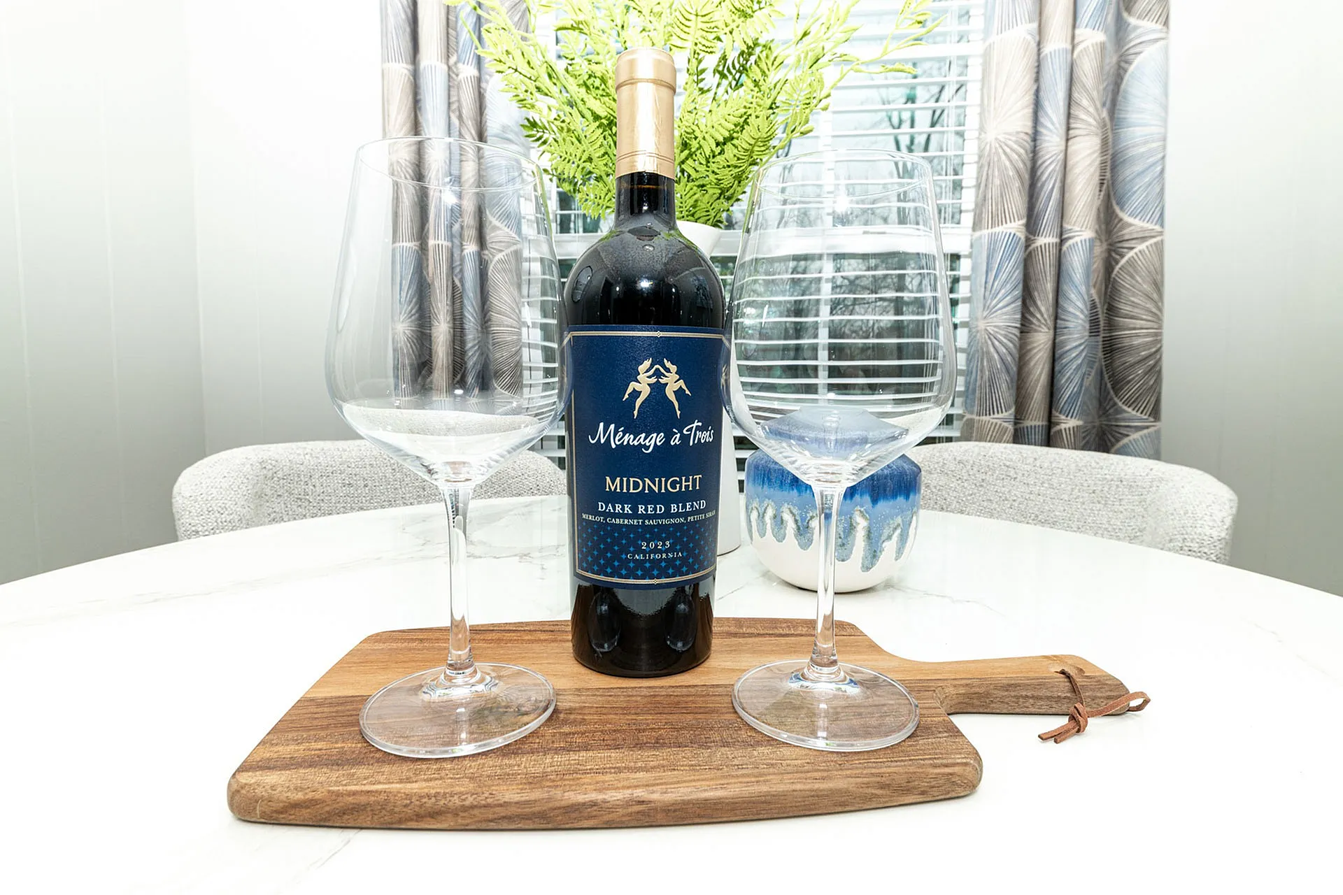 Elegant dining setup featuring a bottle of red wine and two glasses on a stylish wooden platter. Cozy and sophisticated ambiance with modern decor and lush greenery.