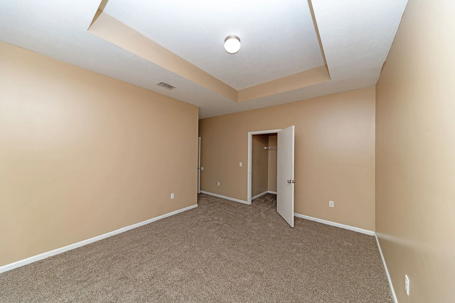 Spacious bedroom with elegant tray ceiling and plush carpet, featuring a walk-in closet and neutral wall tones.