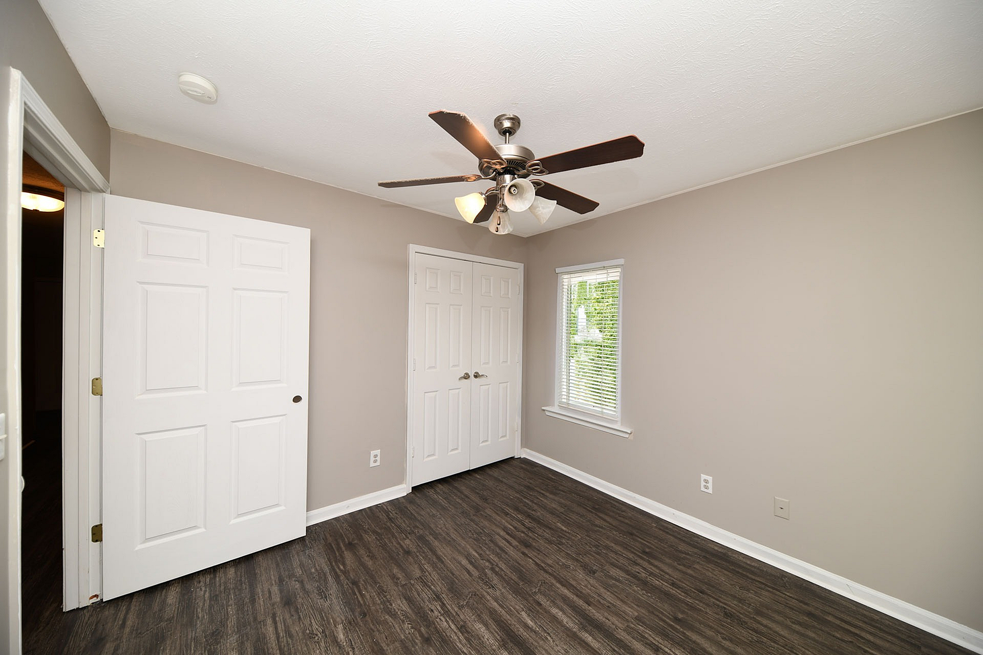 Modern bedroom with sleek wood floors, neutral walls, and ceiling fan, offering ample natural light through a large window.