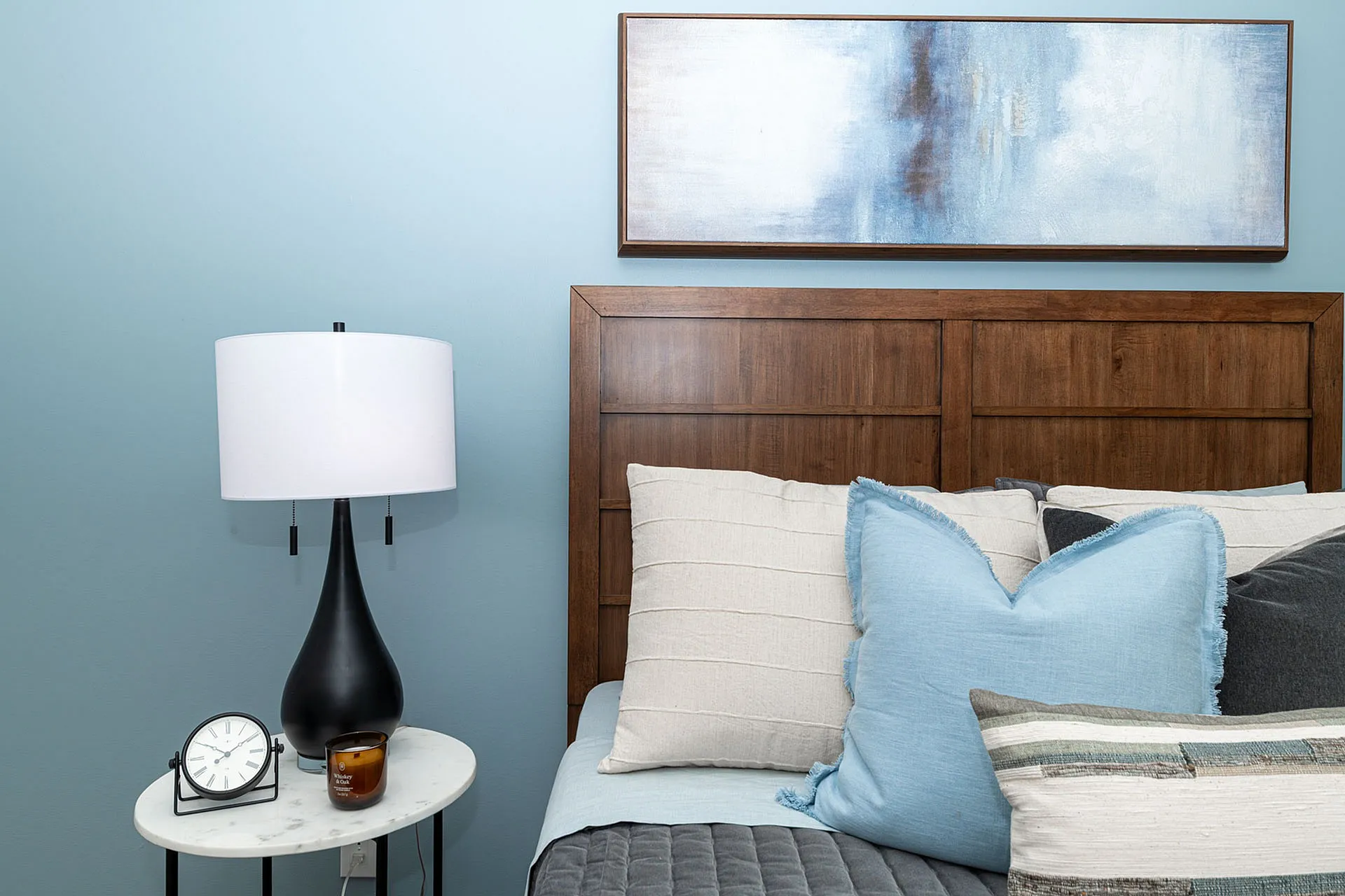 Elegant bedroom with a cozy wooden headboard, soft blue and gray pillows, and a stylish black lamp on a marble side table.