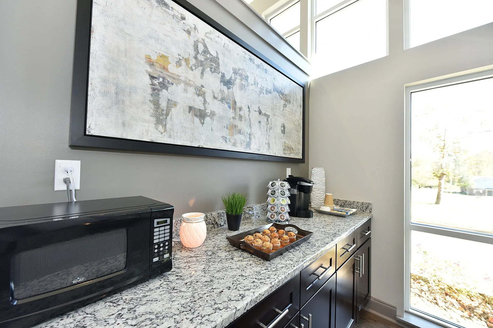 Bright kitchen nook with sleek granite countertop, stylish wooden cabinets, and modern appliances, enhanced by a large abstract painting and natural light from expansive windows.