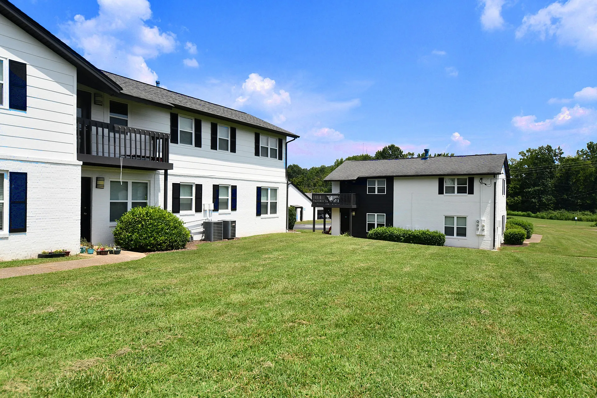 Charming countryside apartment buildings with clean white exteriors, elegant black shutters, and a lush green lawn under a bright blue sky.