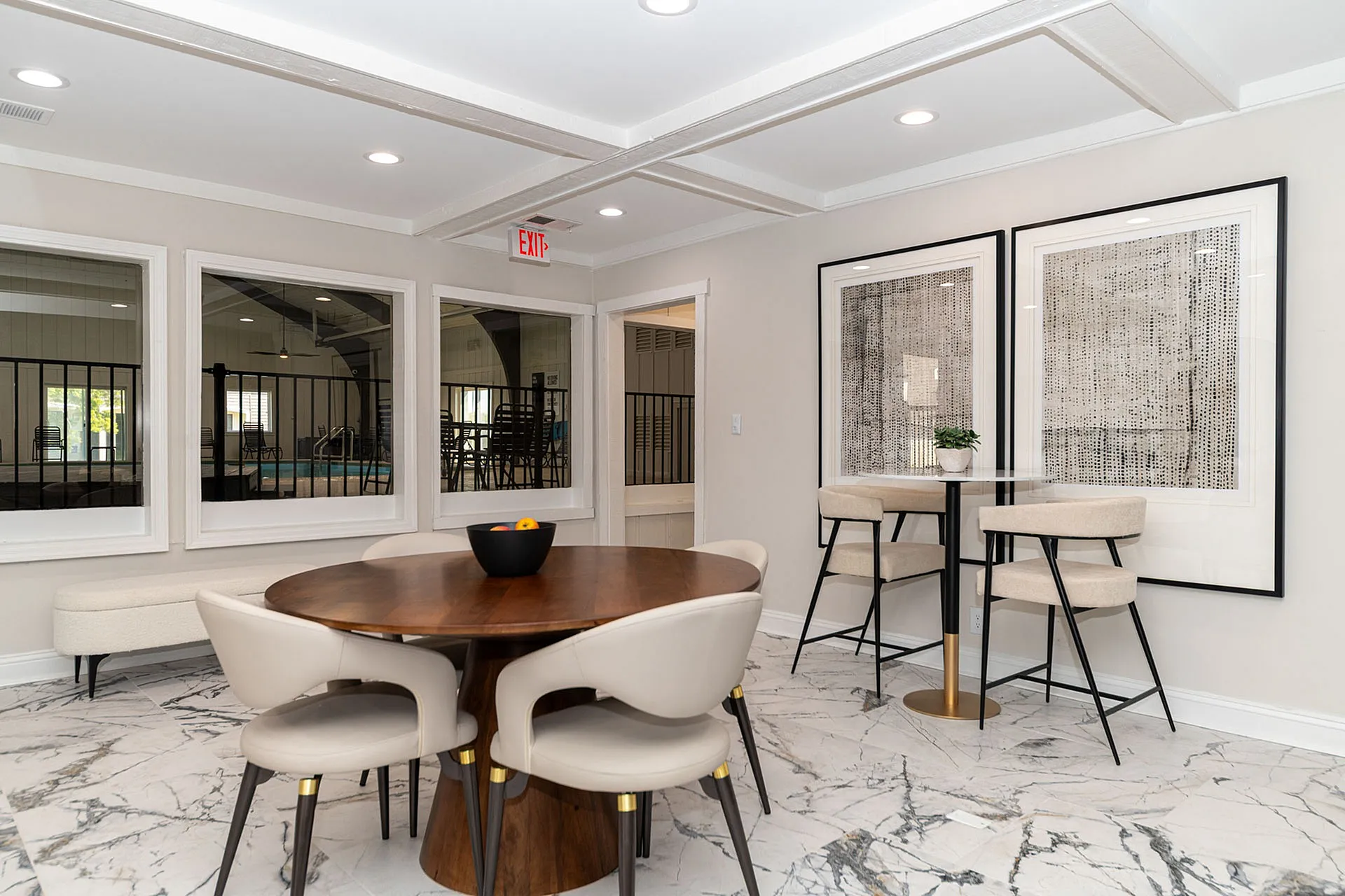 Elegant dining area featuring a round wooden table, modern leather chairs, and stylish wall art with views of an indoor pool through large windows.