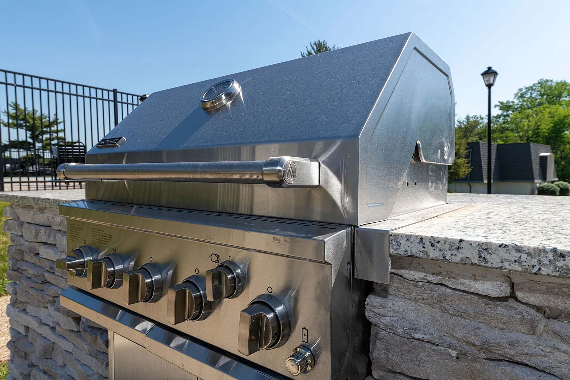 Premium outdoor stainless steel grill with granite countertop, set against a sunny, fenced backyard.