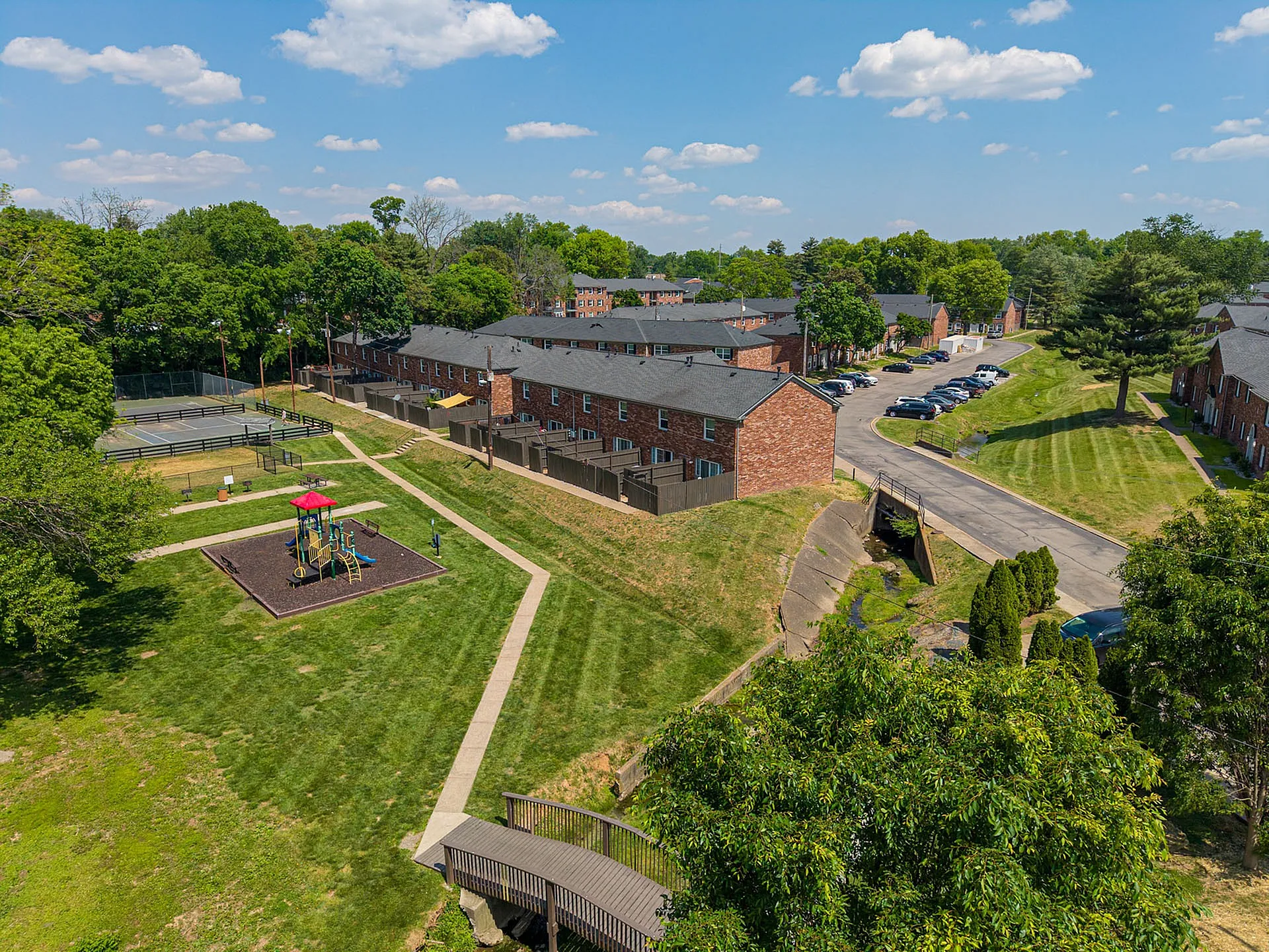Charming aerial view of a suburban apartment complex featuring red brick buildings, lush green lawns, a children’s playground, and tennis courts under a sunny sky.