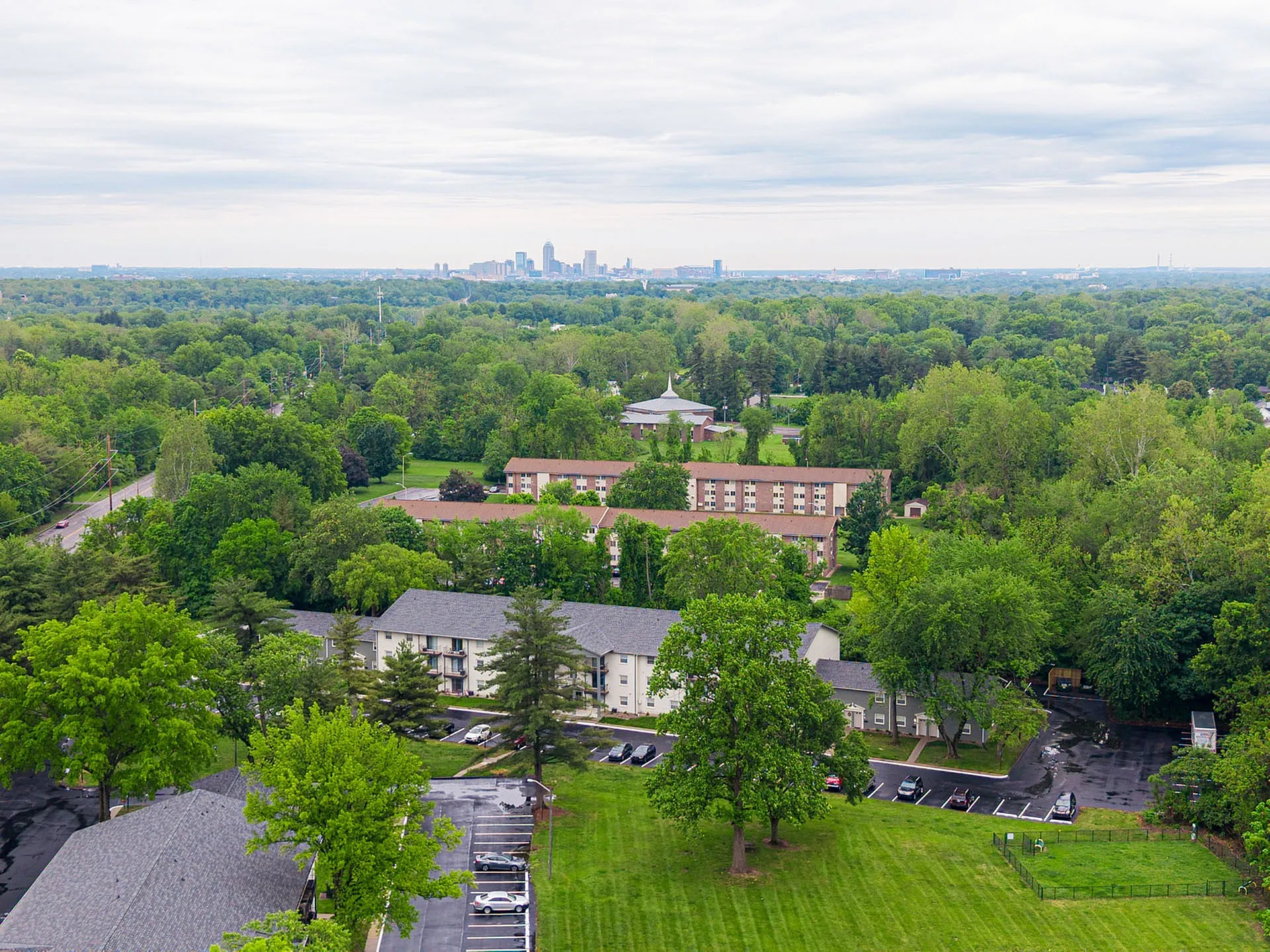 Aerial view of a lush green suburban residential area, featuring modern apartment buildings surrounded by expansive trees and a distant city skyline.