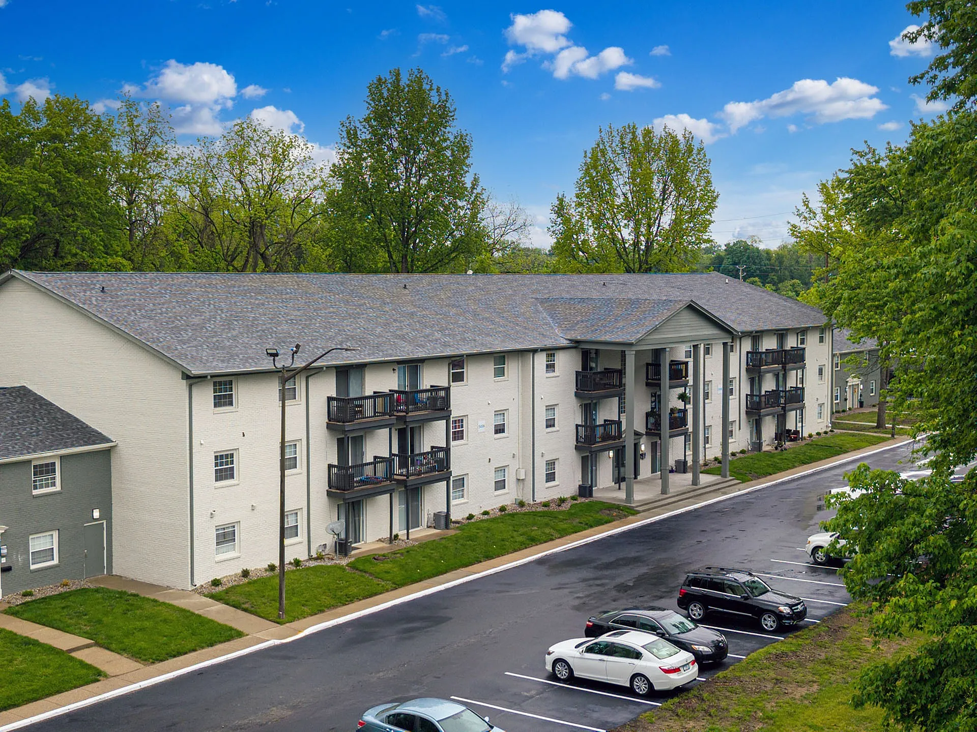 Charming apartment building nestled among lush trees, featuring spacious balconies and ample parking, under a brilliant blue sky.