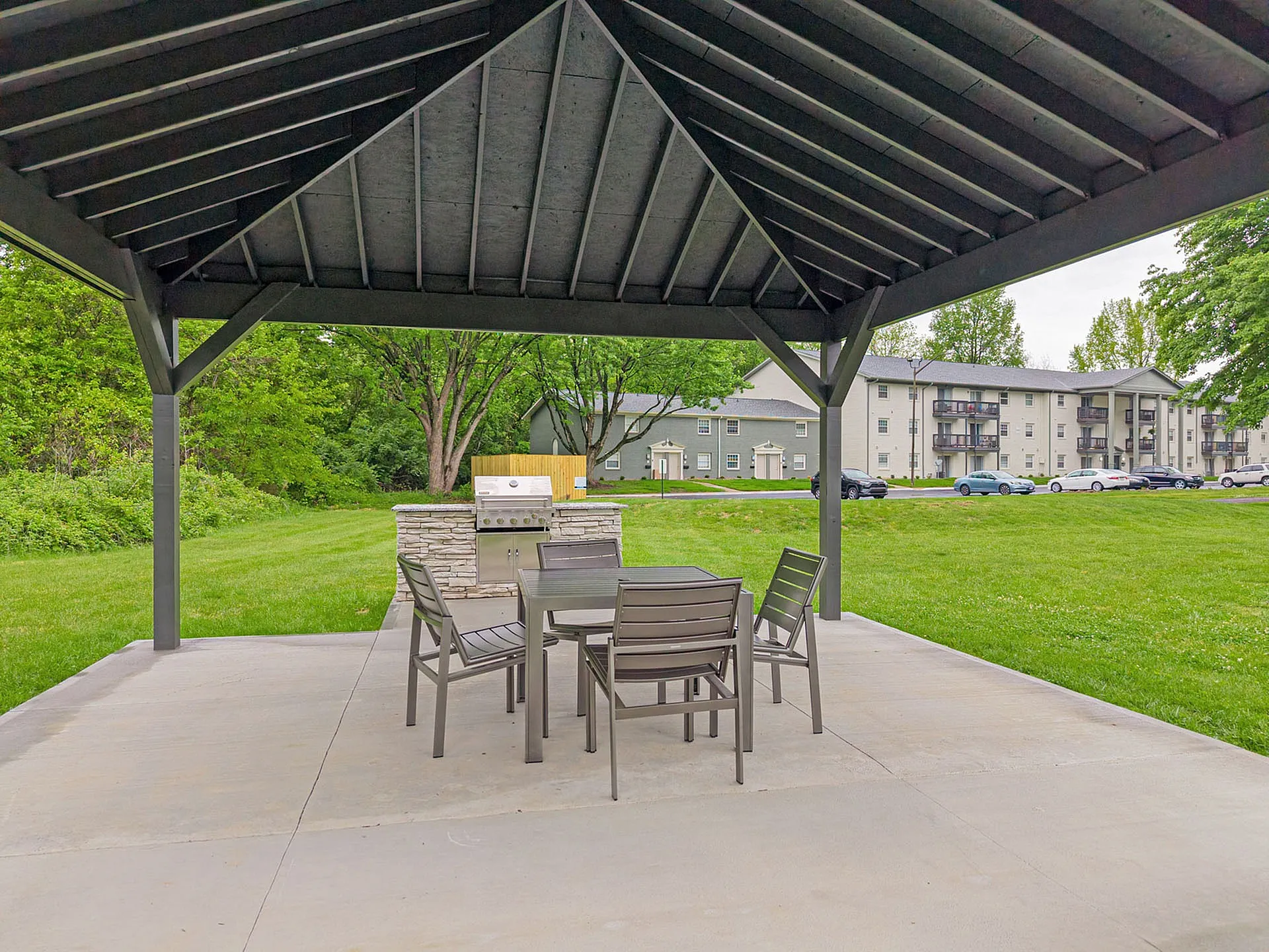 Inviting outdoor pavilion with modern seating and a barbecue grill, set against lush green lawns and charming apartment buildings in the background.