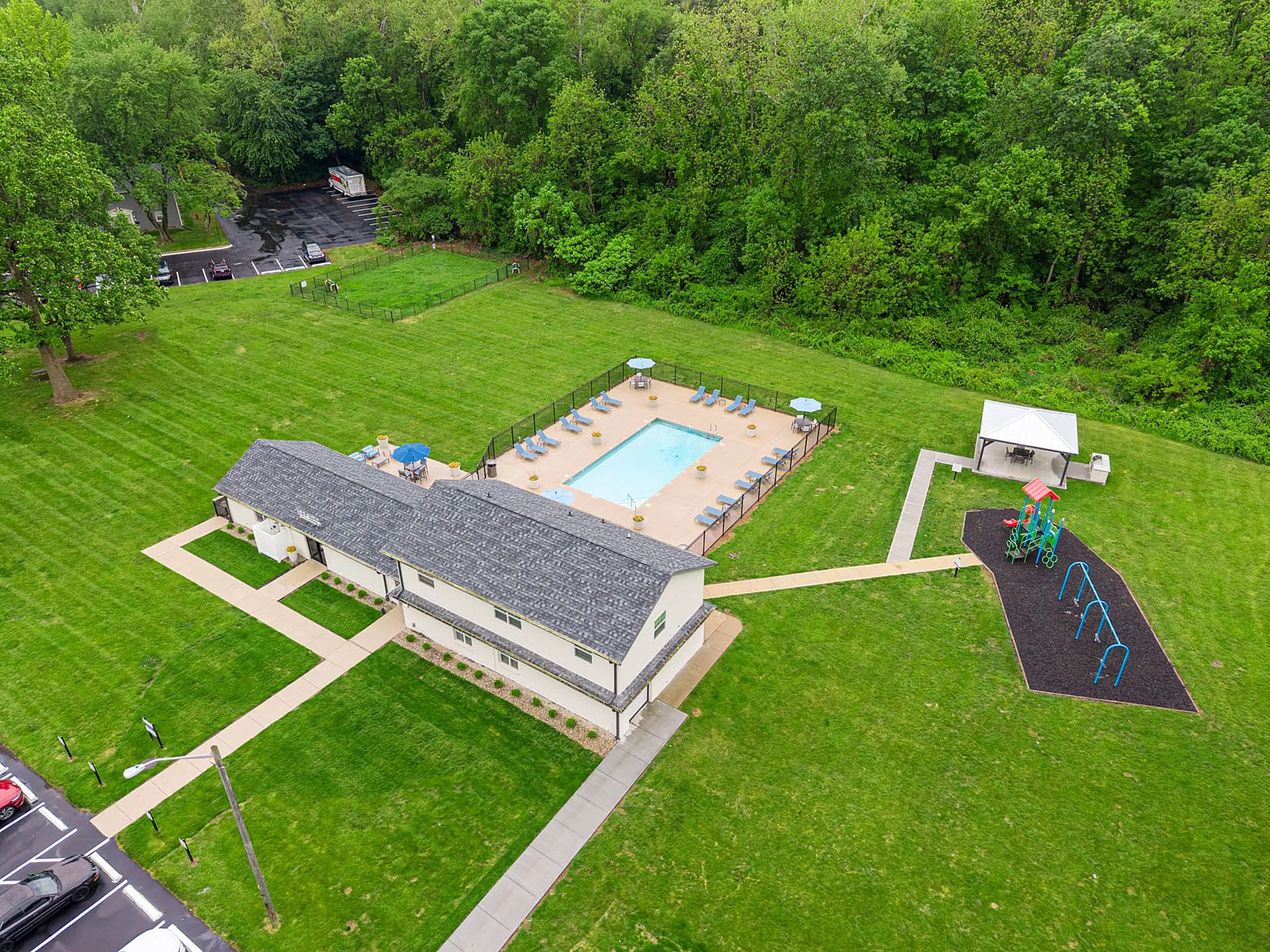 Aerial view of a picturesque apartment complex with a sparkling pool, lush green lawn, playground, and picnic area surrounded by trees.