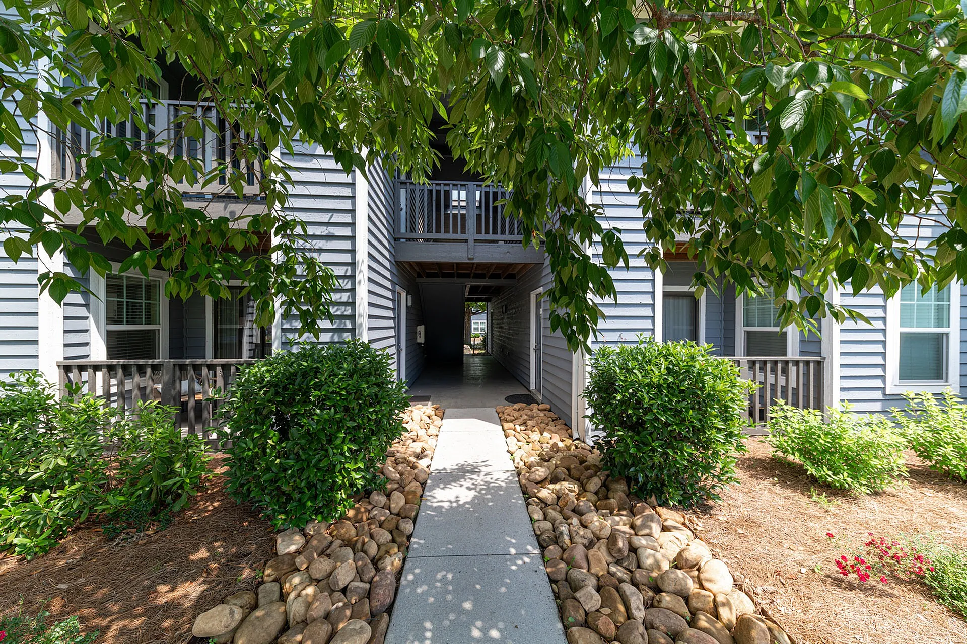 Charming apartment exterior with lush green landscaping, stone pathway, and cozy balconies.