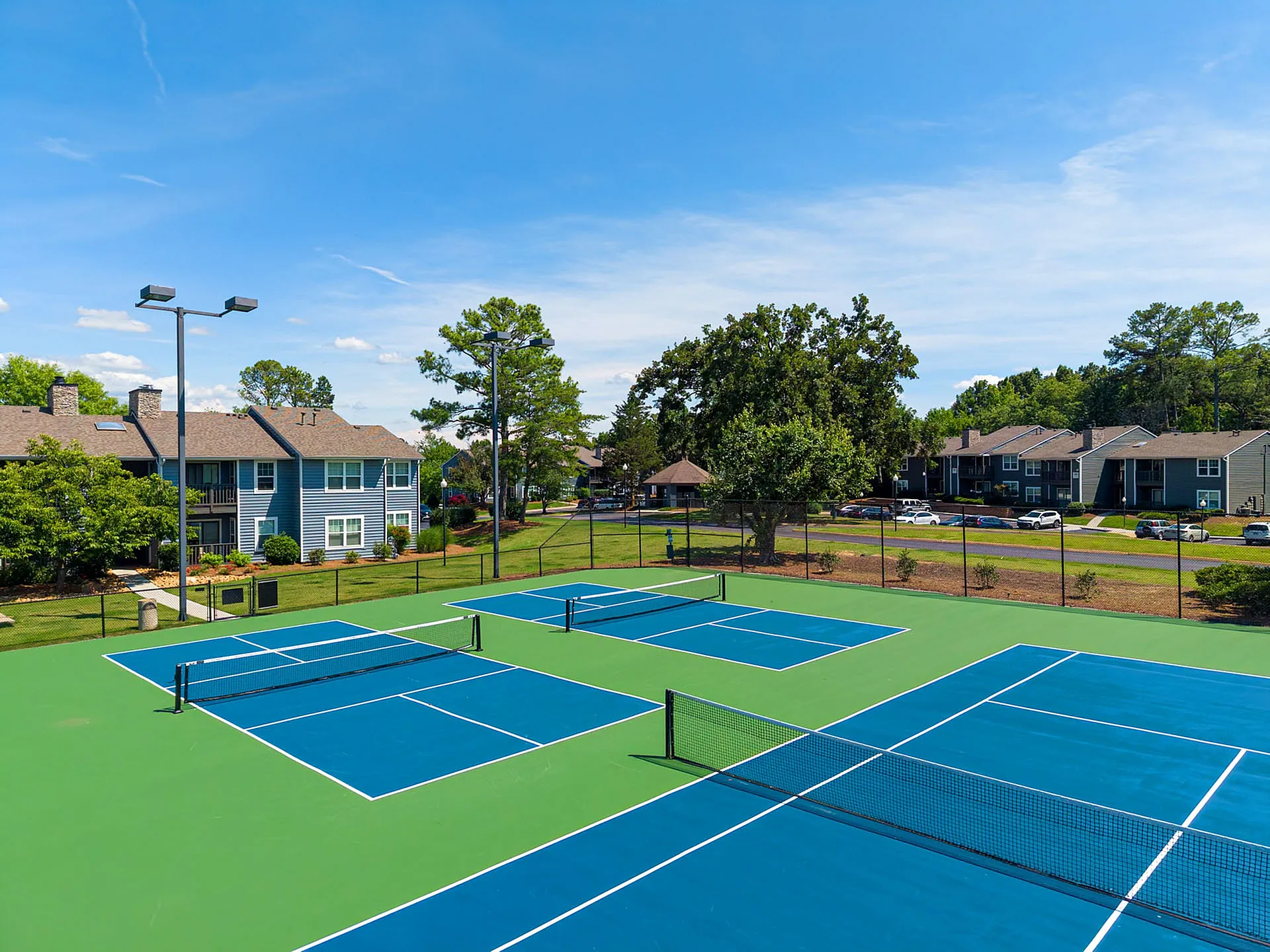 Inviting community view of vibrant tennis courts surrounded by lush landscaping and charming residential buildings under a bright blue sky.