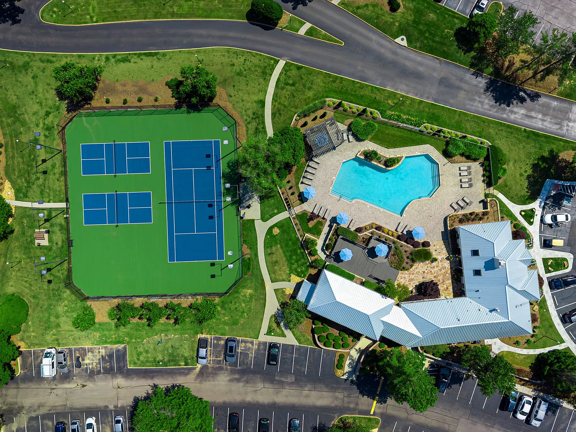 Aerial view of a vibrant apartment complex featuring two tennis courts, a stylish swimming pool with lounge chairs, and modern facilities surrounded by lush greenery.