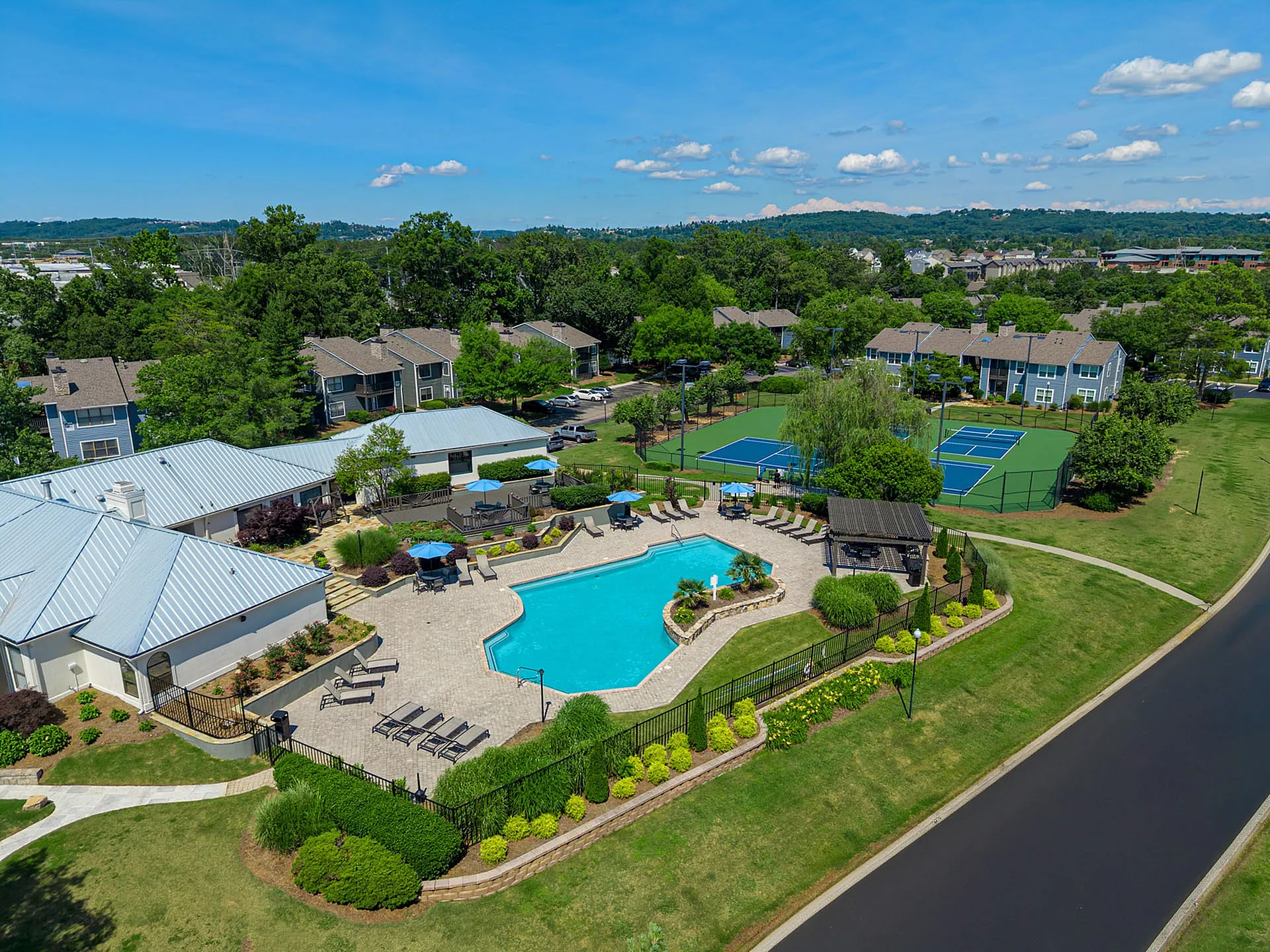 Luxurious apartment community with a sparkling pool, lush greenery, and multiple tennis courts under a clear blue sky.