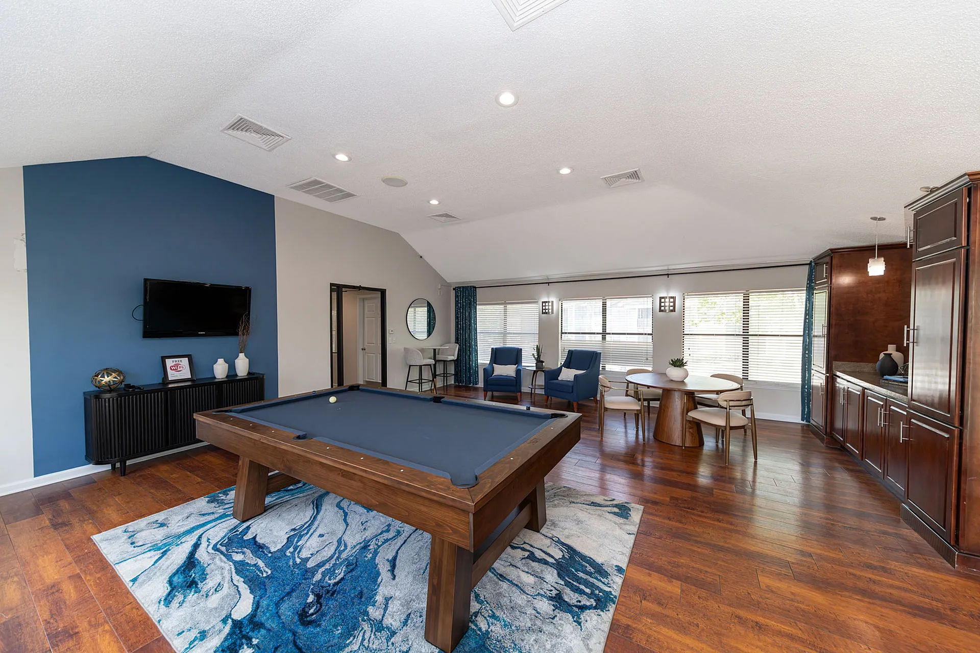 Elegant community lounge featuring a sleek billiards table, cozy seating area, and bright dining space under large windows.