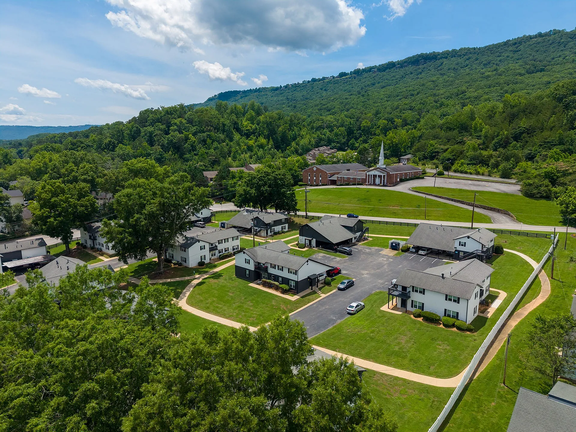 Charming aerial view of a tranquil apartment community nestled in lush greenery with convenient pathway access and a picturesque backdrop of rolling hills and a church.