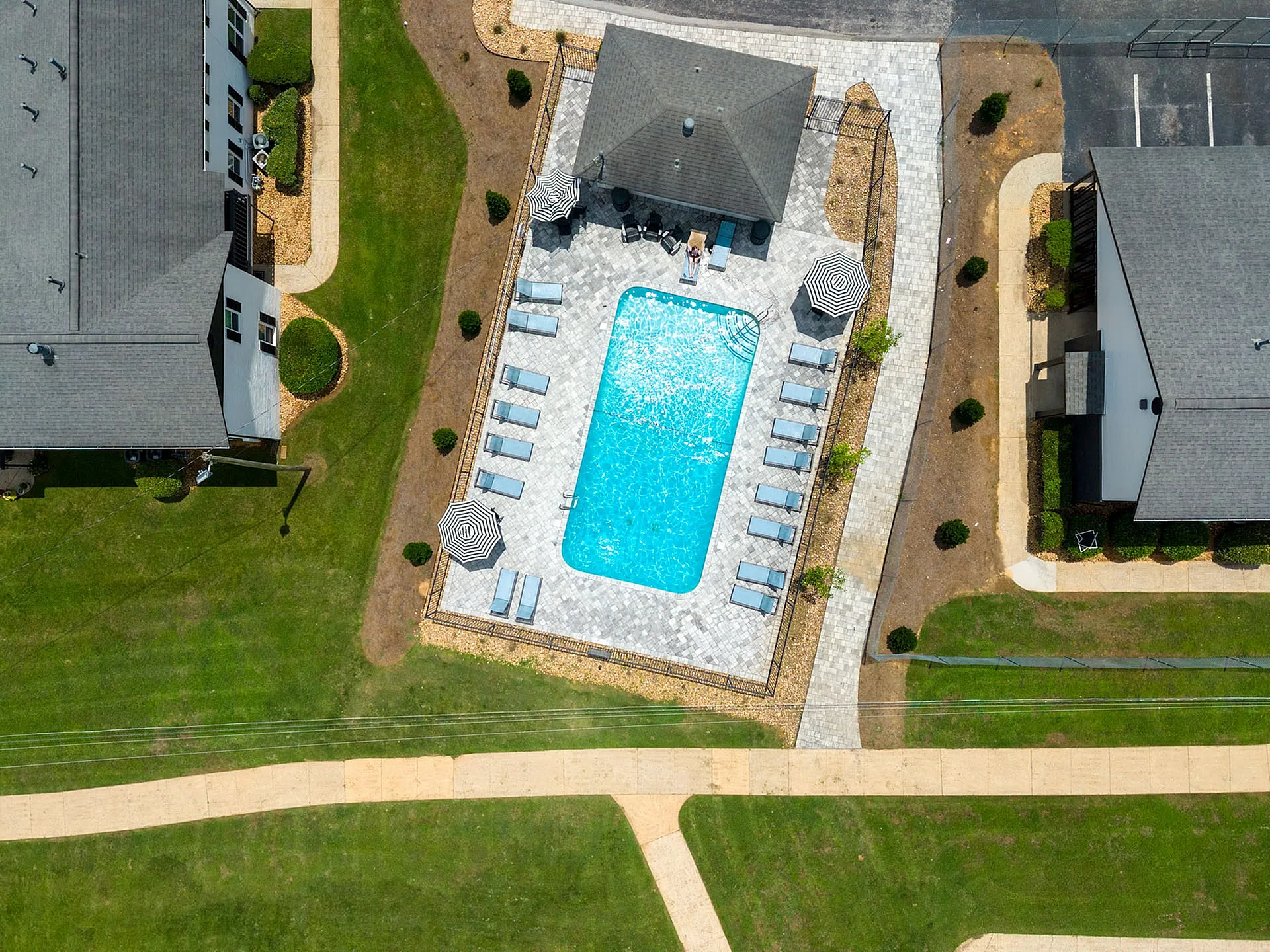 Aerial view of a luxurious apartment complex featuring a sparkling swimming pool with sun loungers and umbrellas, surrounded by well-manicured lawns and walking paths.