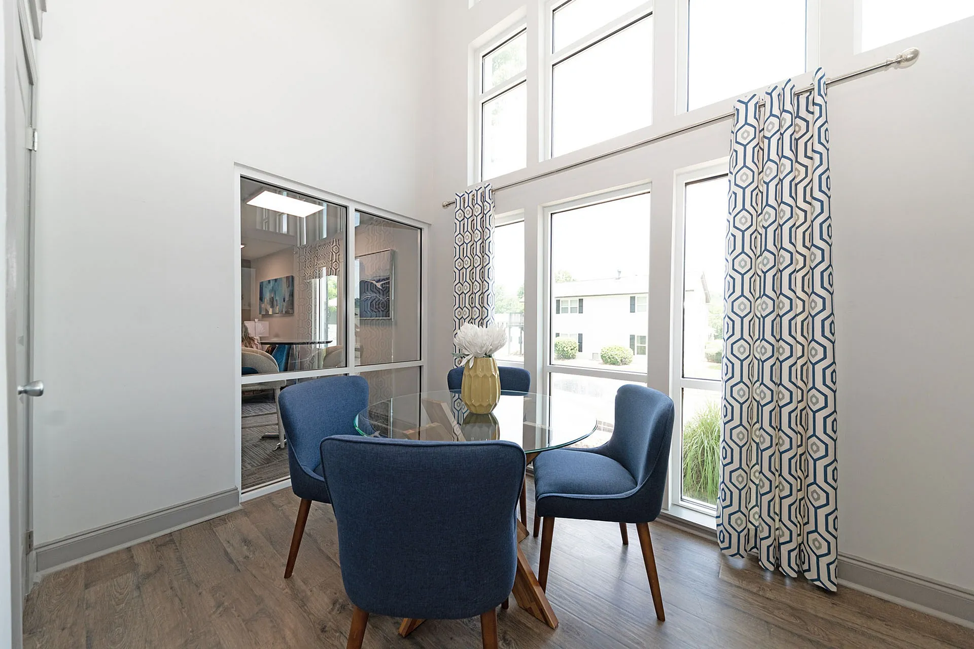 Bright dining area with modern blue chairs around a glass table, set against floor-to-ceiling windows and stylish geometric curtains.