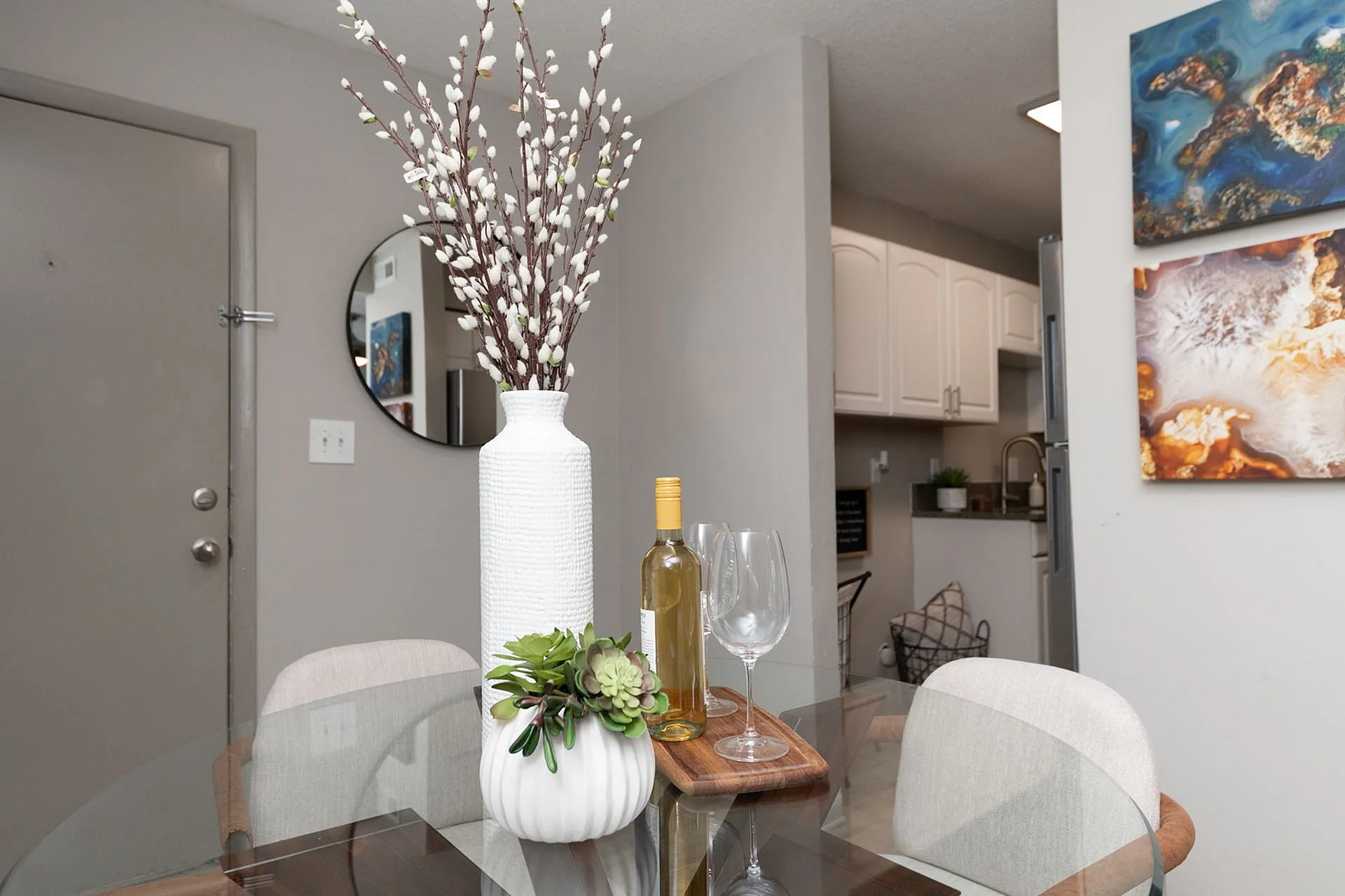 Modern dining area with elegant glass table, chic white vase, and artistic wall decor, adjacent to a stylish kitchen.